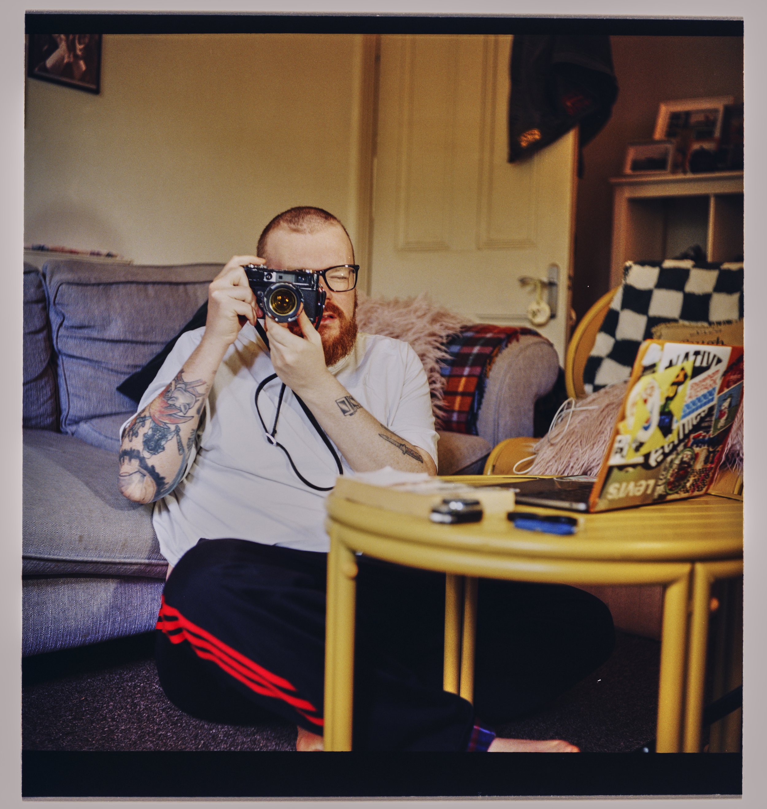 A person with short hair and tattoos on their arms is sitting on the floor taking a selfie in a mirror, with a camera in their hand and their face partially visible.