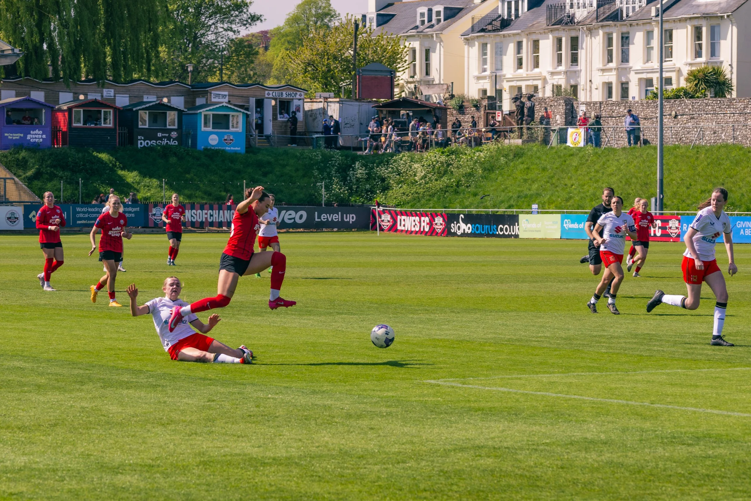 Women playing soccer on a green field during a sunny day, with spectators and houses on a hill in the background.