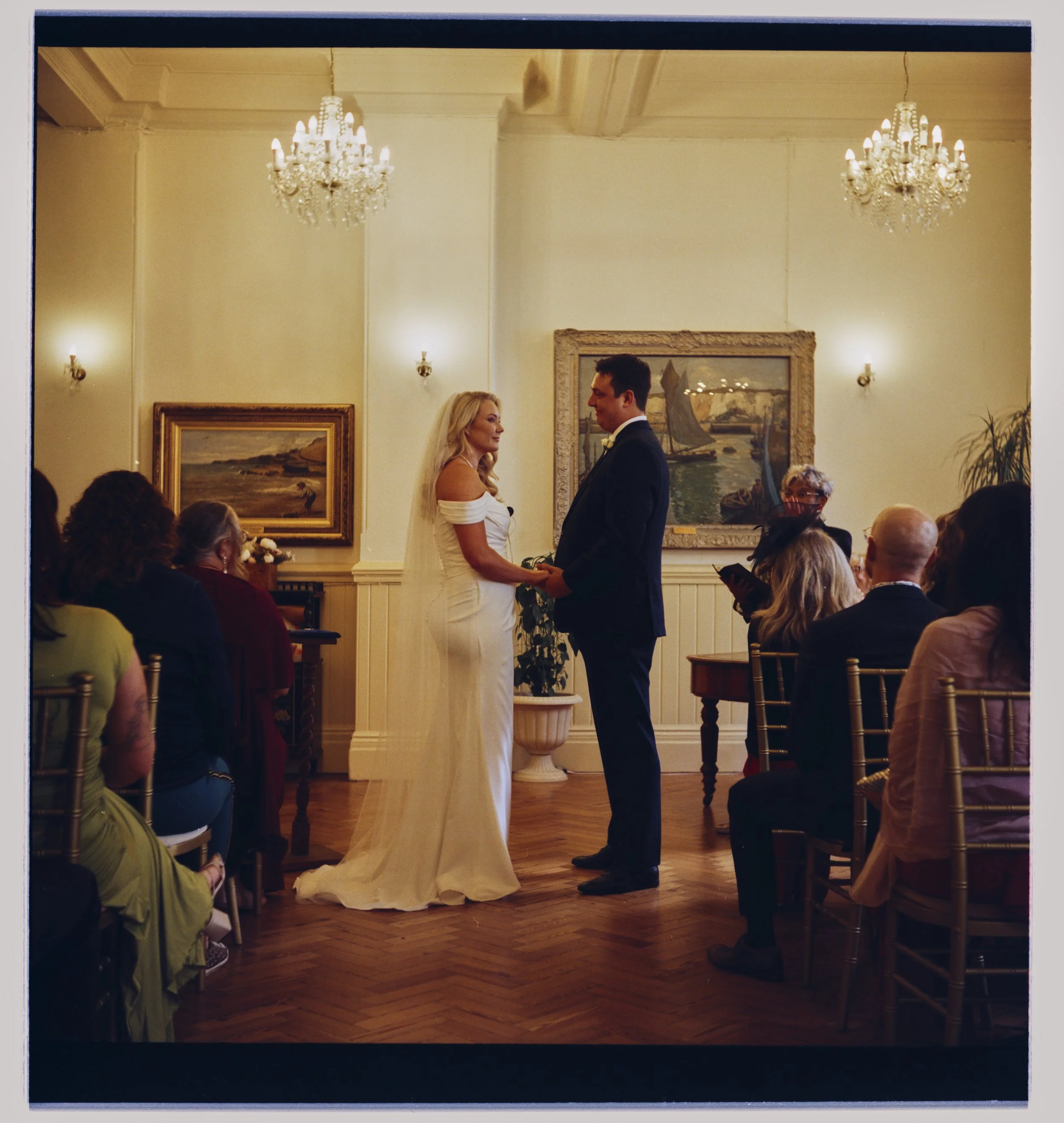 A wedding ceremony taking place in an elegant room with chandeliers, paintings on the wall, and guests seated on chairs. The bride and groom are standing and holding hands, facing each other.