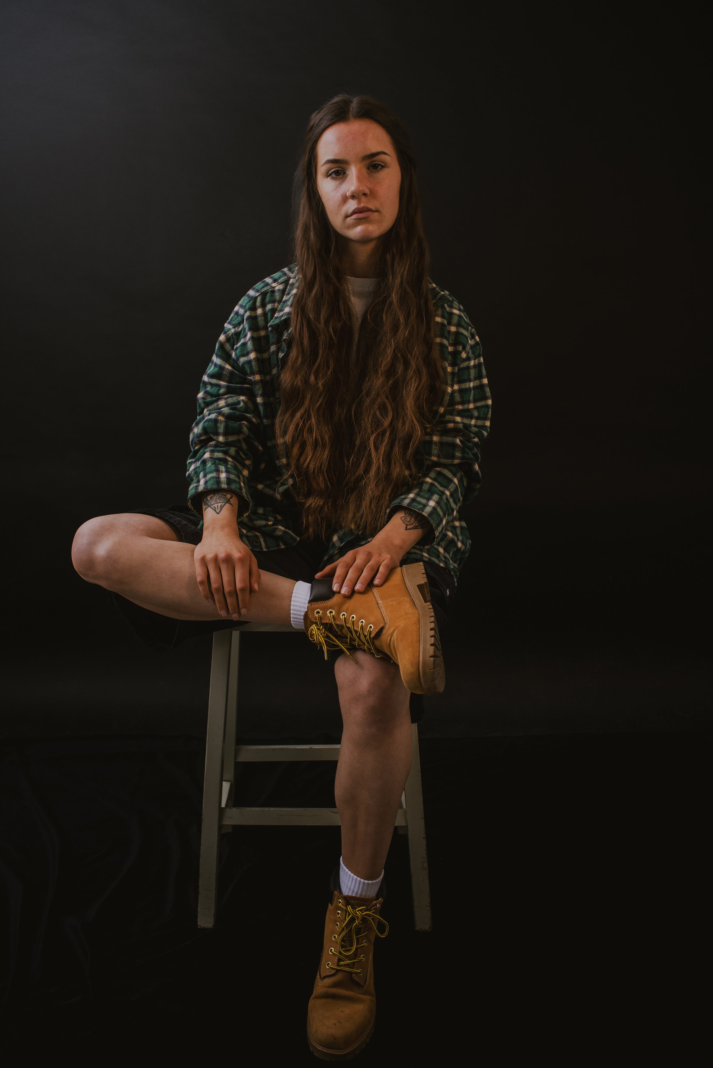 A young woman with long wavy brown hair, wearing a green and black plaid shirt, sitting on a stool against a black background. She has tattoos on her arms, is wearing white socks, tan work boots, and black shorts. She is looking directly at the camer