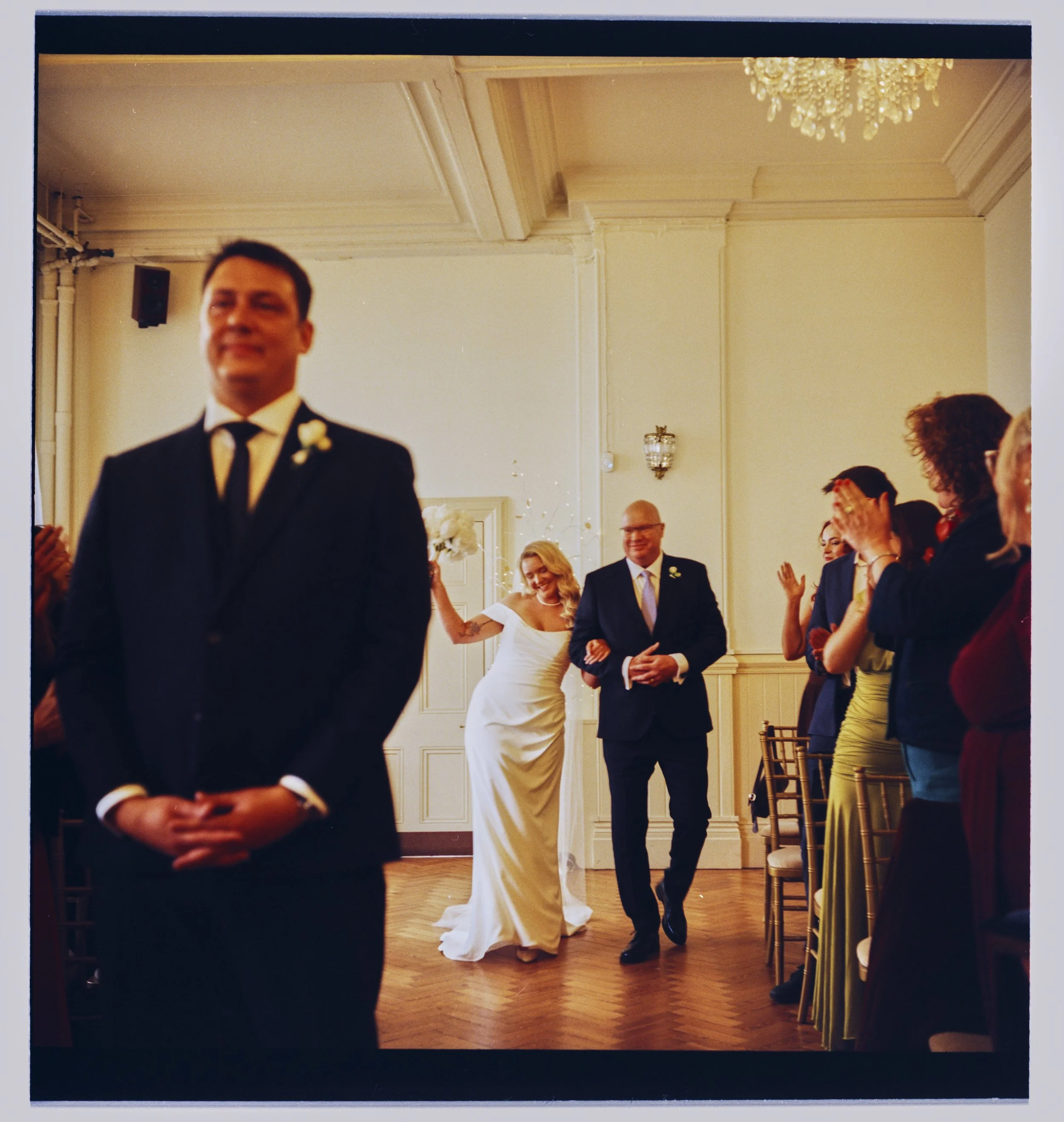 A wedding reception with guests standing and clapping, a bride in a white gown, and a groom in a black suit, in a decorated elegant room with a chandelier and ornate wall details.