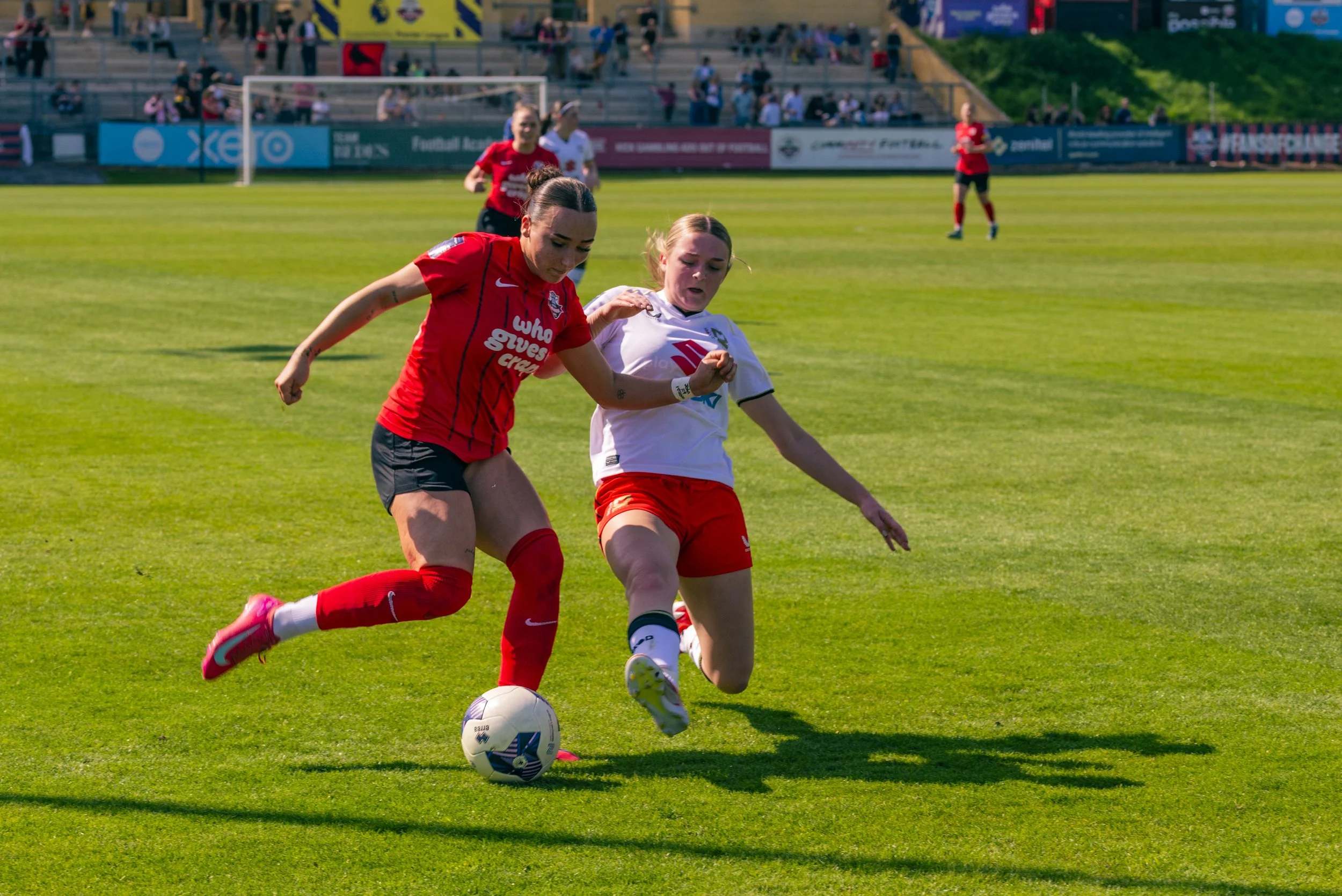 Two female soccer players racing for the ball on a grass field, with spectators in the stands in the background.