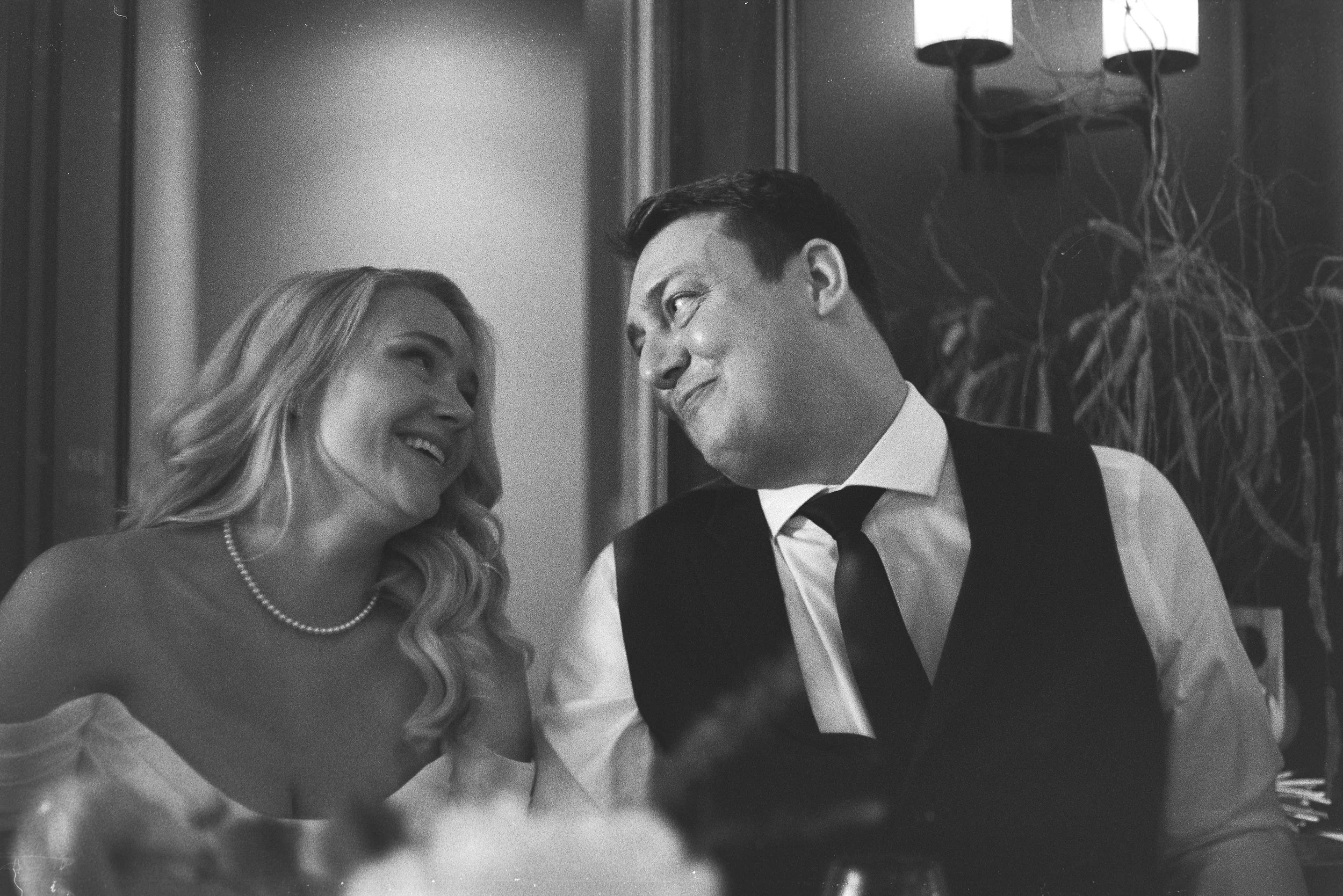 Black and white photo of a smiling woman in a wedding dress with pearl necklace and a man in a tuxedo, both looking at each other, at a formal event or wedding celebration.