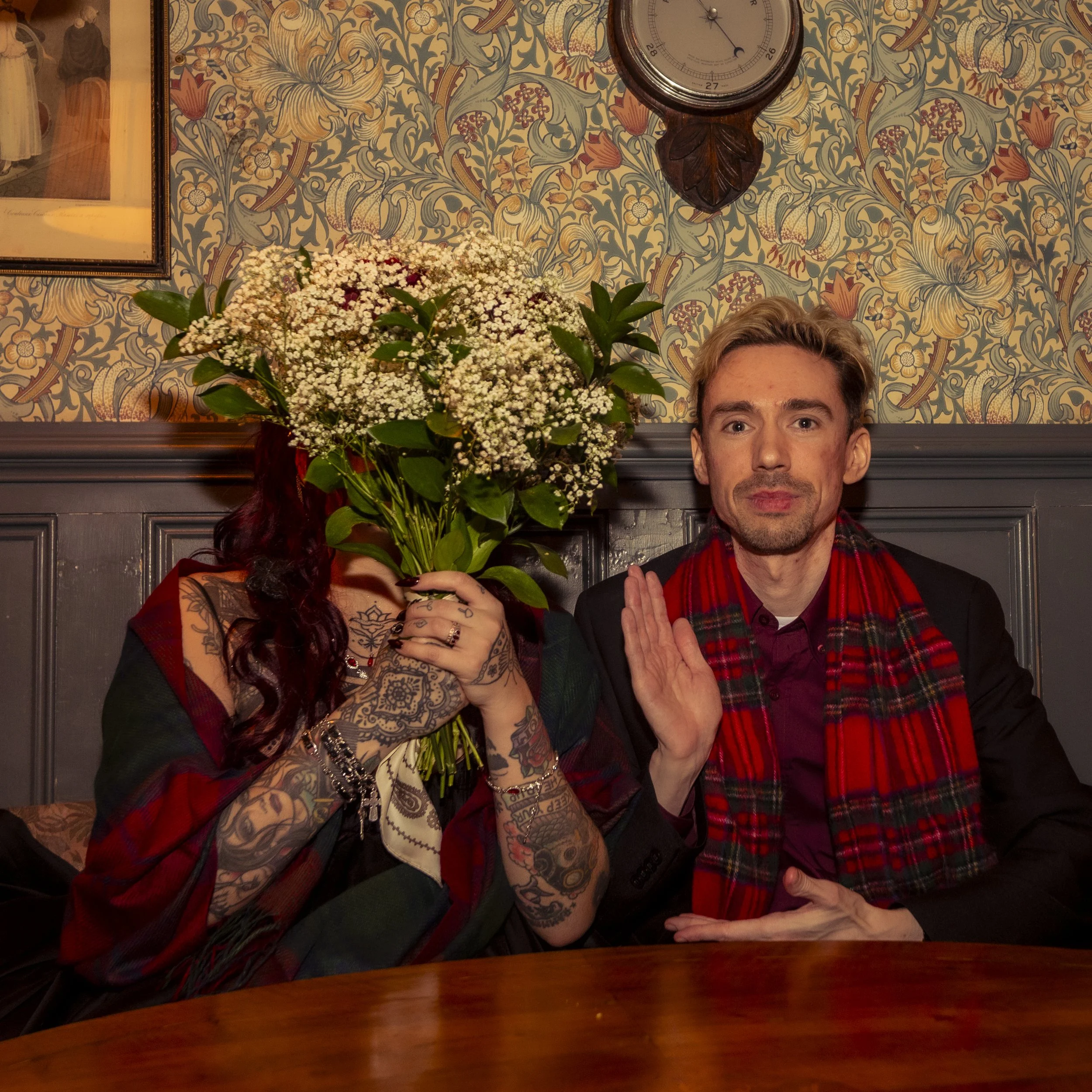 A man with short hair and a woman with tattoos holding white flowers seated at a wooden table with a patterned wall, clock, and picture in the background.