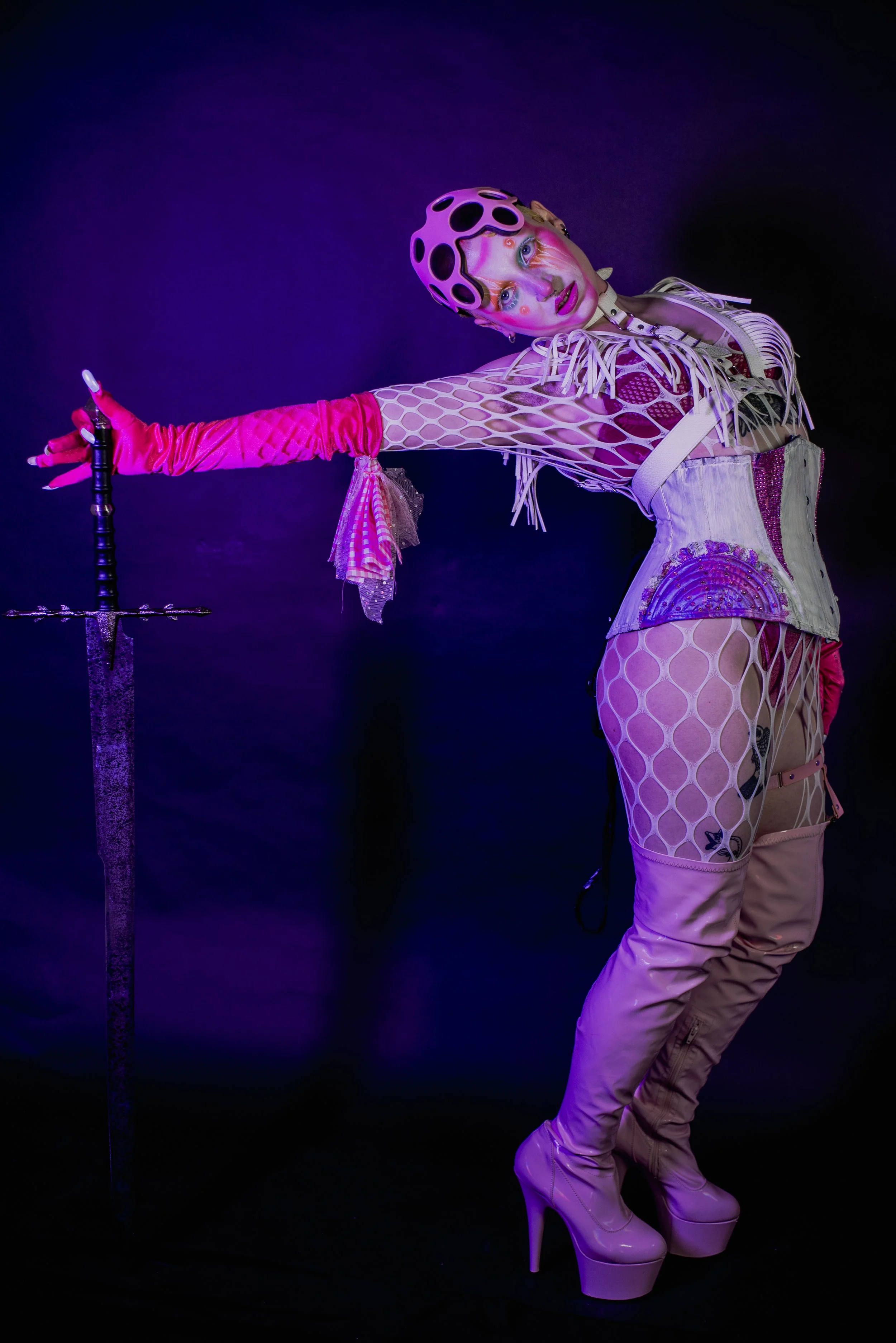 Person dressed in futuristic, pink and purple fashion with platform boots and fishnet stockings, holding a sword against a dark backdrop with purple lighting.