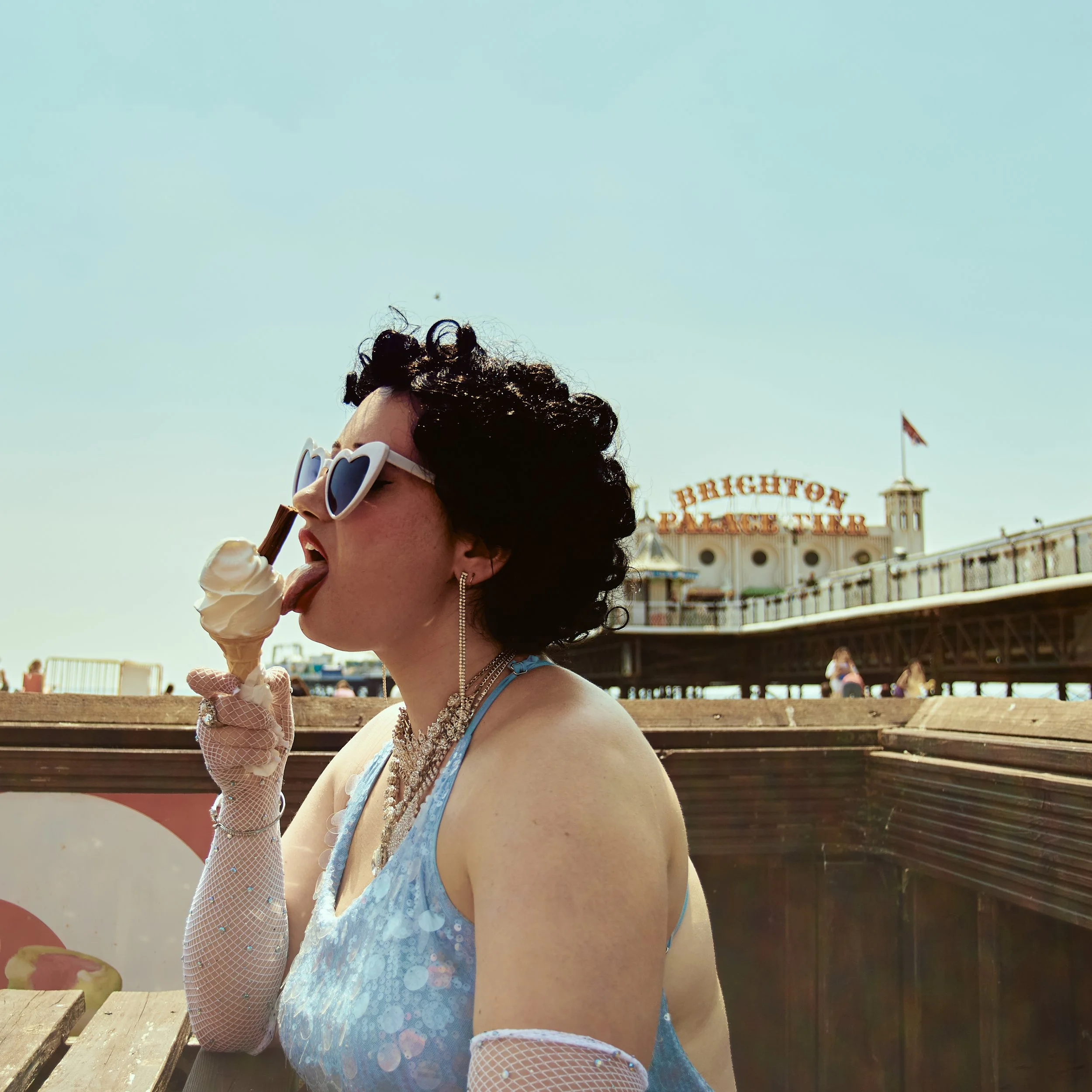 A woman wearing white heart-shaped sunglasses, a blue dress with sequin detailing, and fishnet gloves is eating ice cream in an outdoor amusement park setting with a sign in the background that says 'Brighton Pier'.