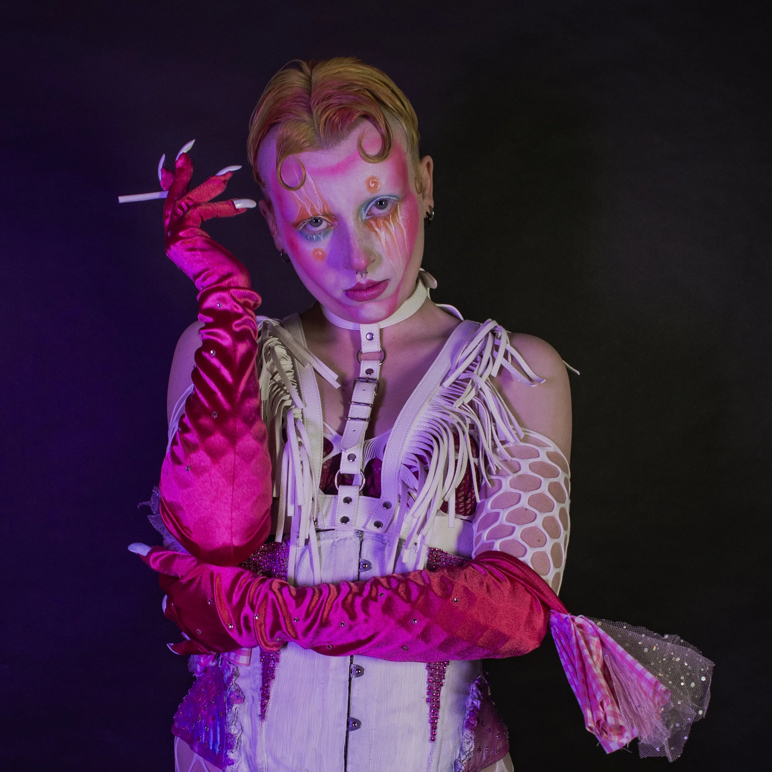 Person with pastel-colored hair, makeup with bright colors, wearing a strappy top with a futuristic design, long pink gloves, and a skirt with sequins and glitter.