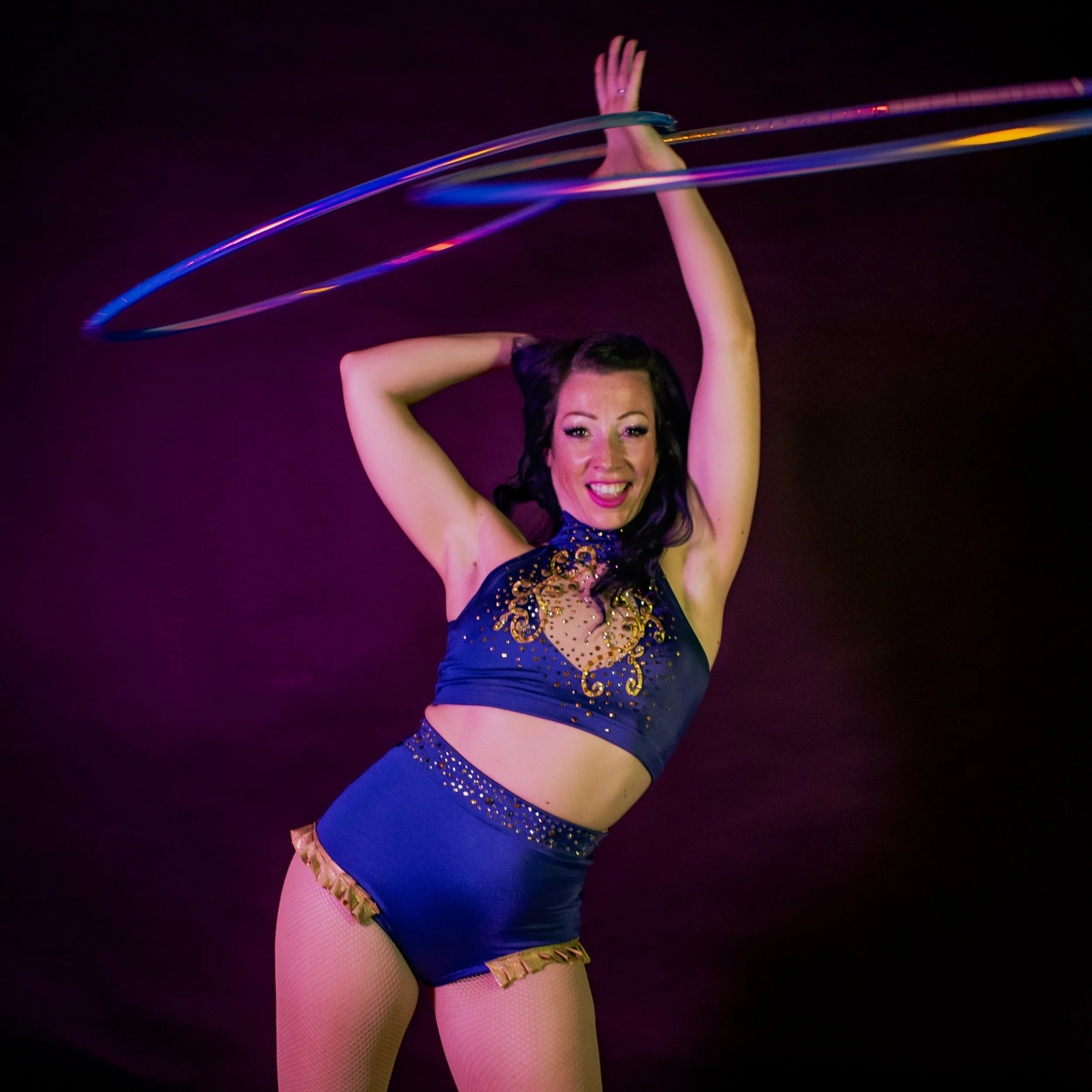 A woman in a shiny blue and gold costume is performing with glow hula hoops, with her face smiling against a dark background.