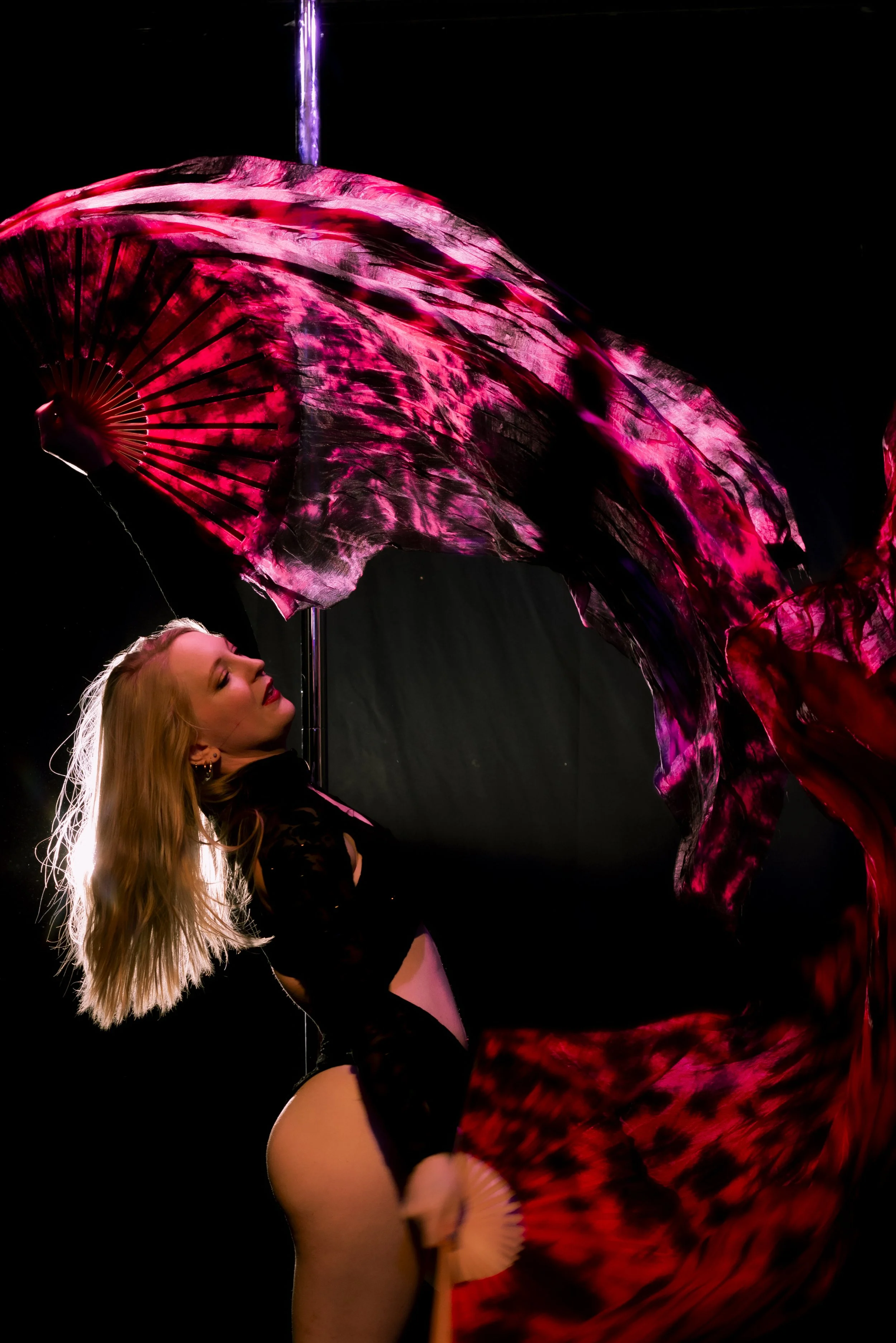 A woman with long blonde hair is performing with a large, colorful, illuminated fan. The fan has pink, black, and red patterns, and the background is dark, highlighting the vibrant colors of the fan.