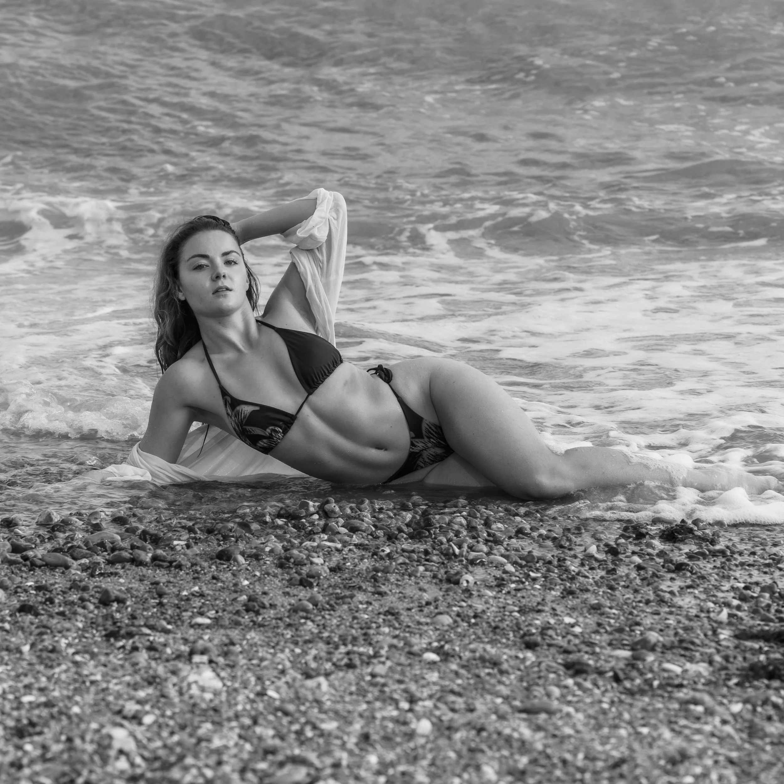 A woman lying on her side on a pebbled beach with her hair loose, wearing a bikini top, with the ocean waves behind her.