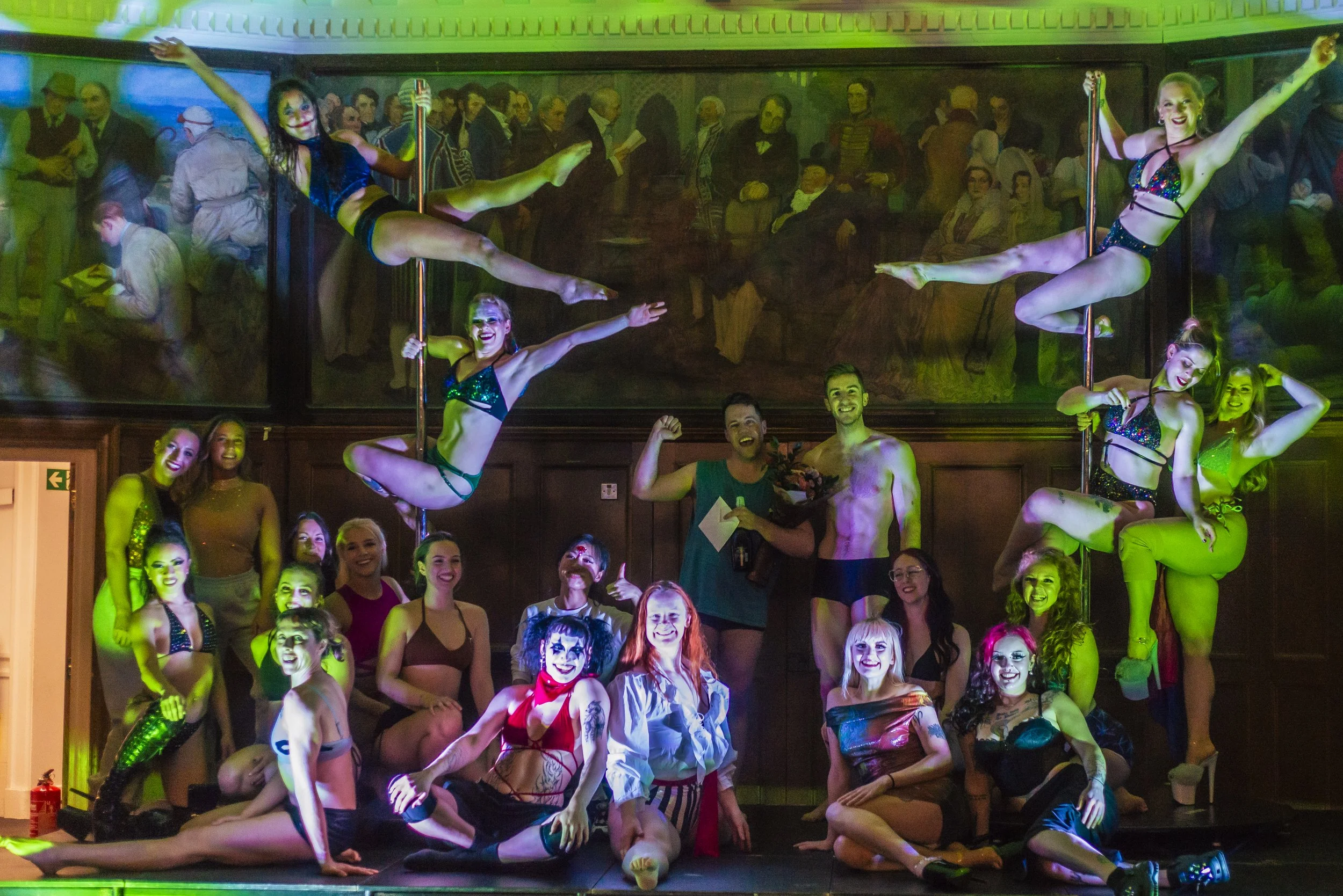 Group of performers and audience members in a circus or club setting, with some performers on poles and others sitting or standing around. The scene is colorful and lively.