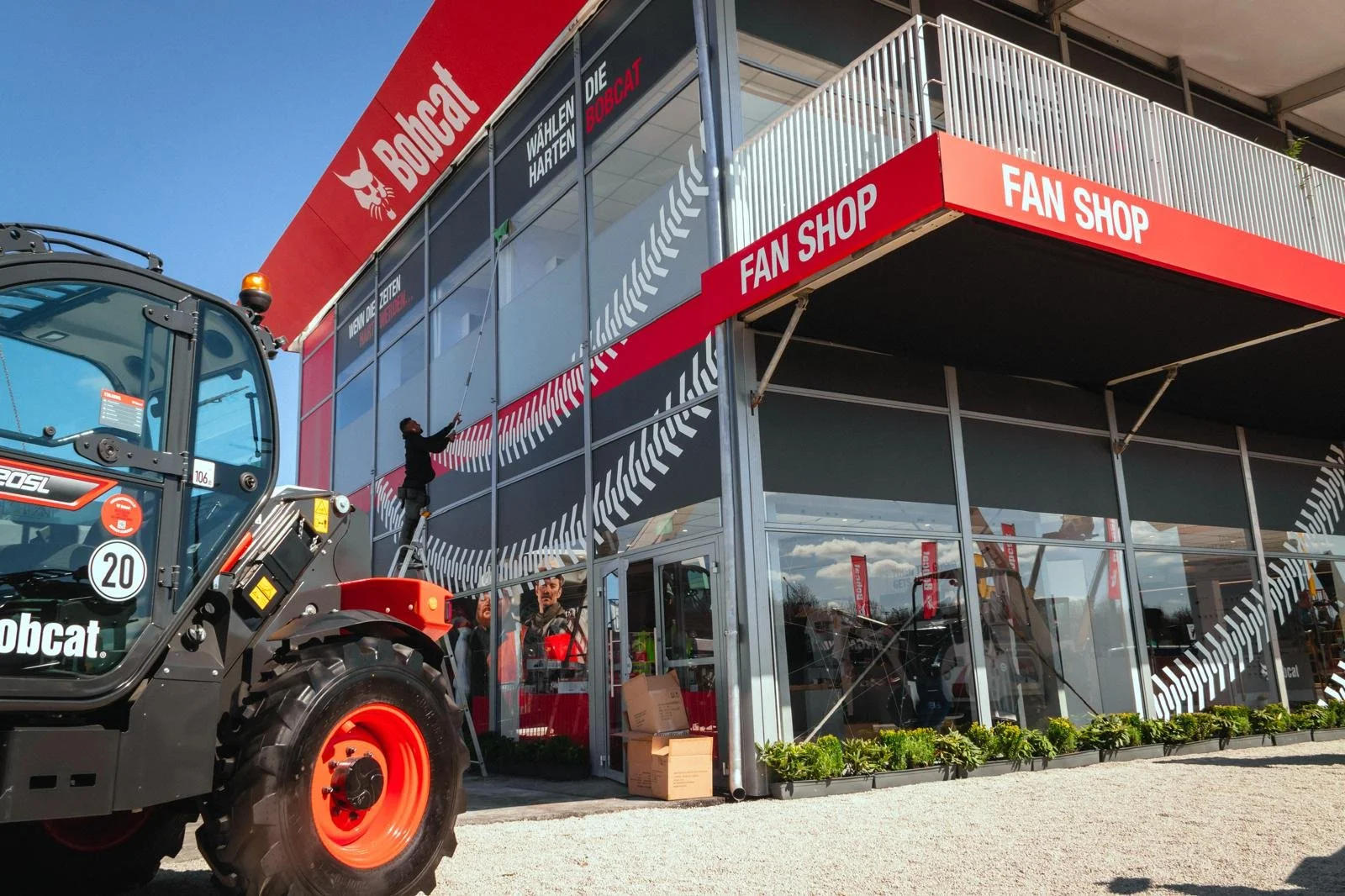 Façade d'un magasin Bobcat, spécialisé dans la vente et la réparation d'équipements de construction, avec un personnel nettoyant les vitres et un tracteur en avant-plan.