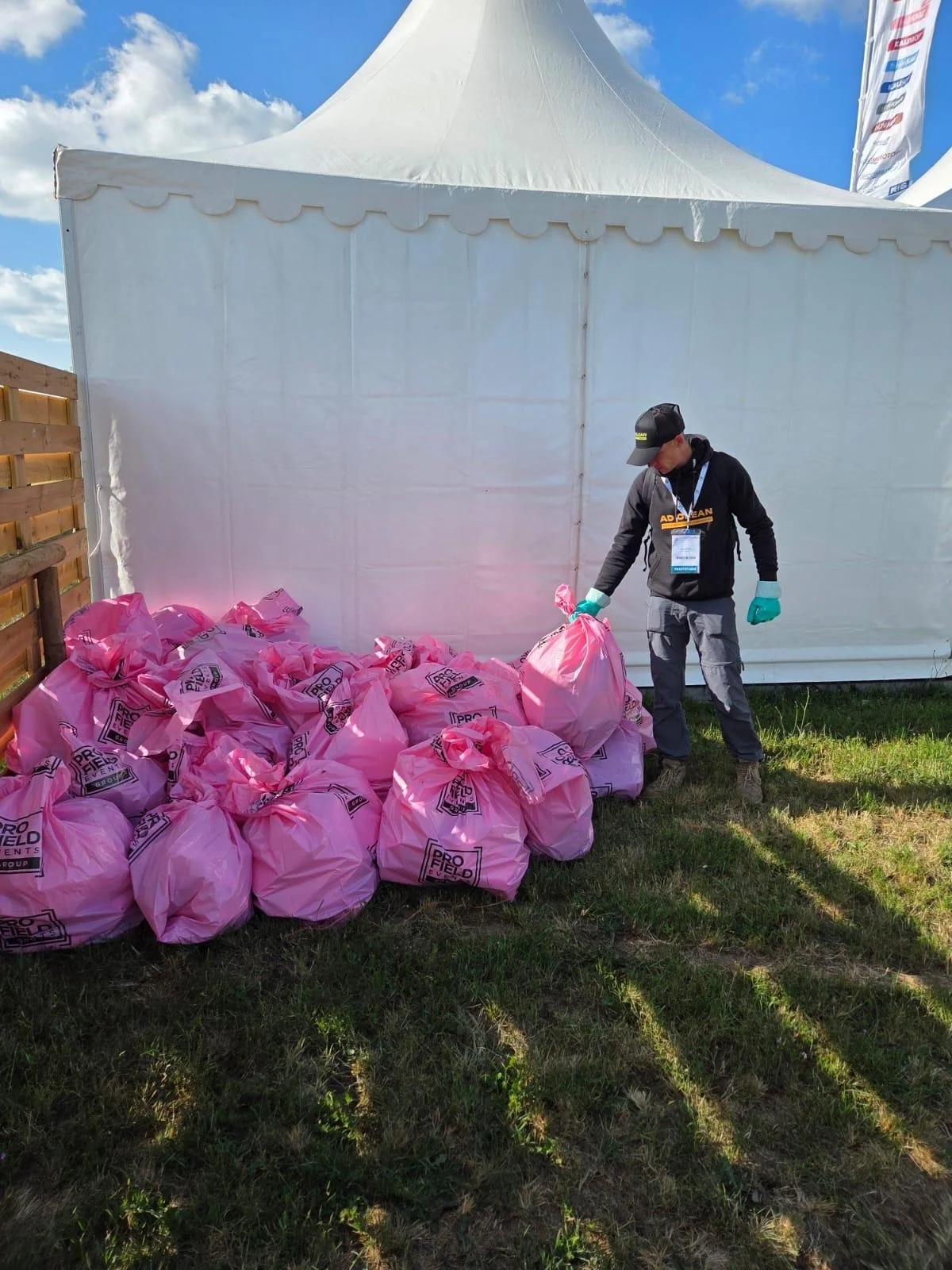 Un employé de l'entreprise AD Clean portant des gants verts ramasse des sacs rose dans un espace extérieur lors d'un événement, avec une tente blanche en arrière-plan et un ciel bleu avec des nuages.