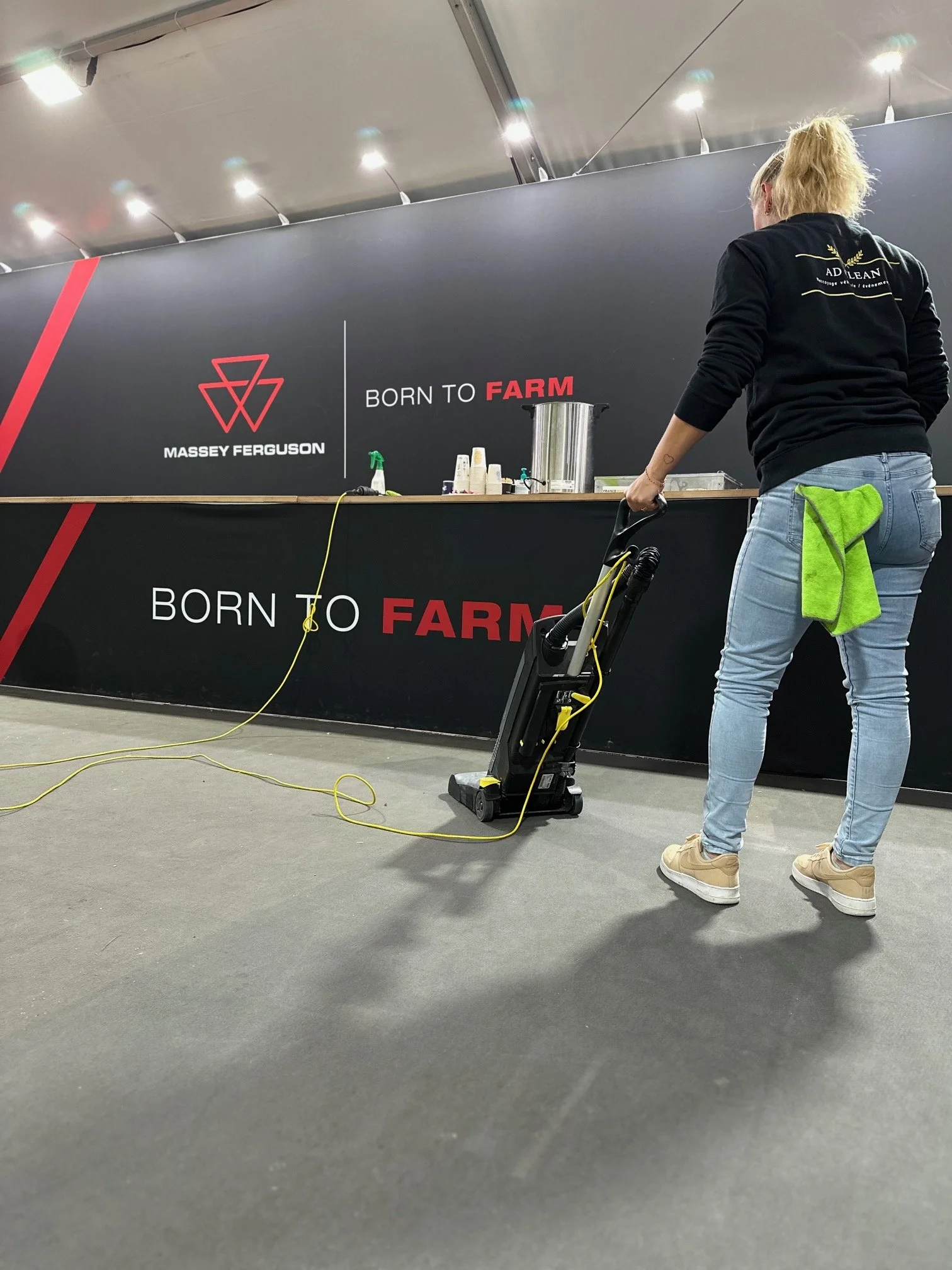 Une employée de AD Clean avec une serviette verte dans la poche de son jean marche près d'une machine dépoussiérante dans un stand de salon,  avec un mur noir derrière affichant le logo de Massey Ferguson et la phrase "Born to Farm".