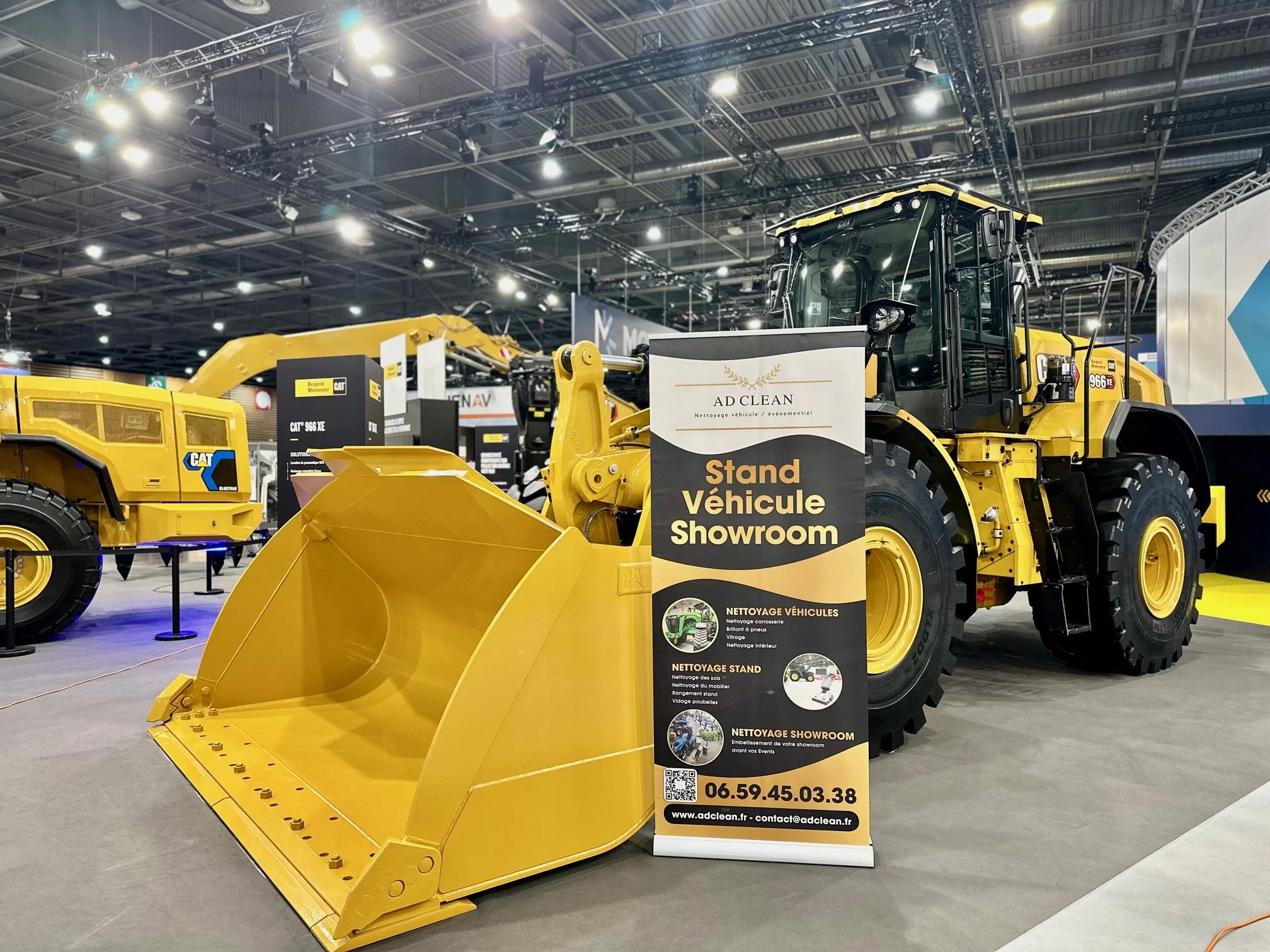 Un grand chargeur de construction jaune exposé dans un hall d'exposition avec une bannière publicitaire pour le prestataire de service AD Clean.