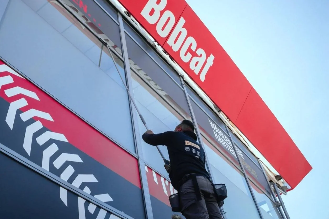Un employé de l'entreprise AD Clean nettoie la façade d'un stand d'exposition avec une échelle, portant une veste noire et un sac à la taille.