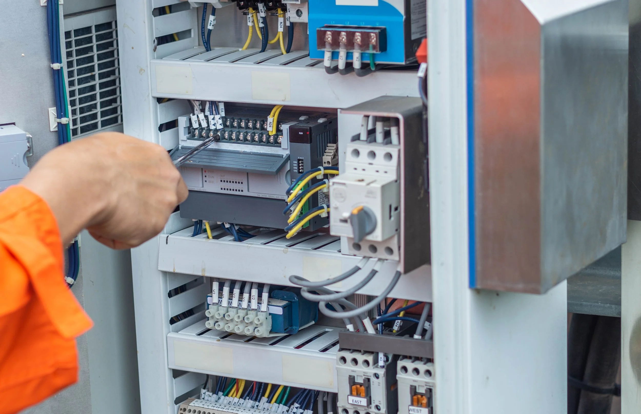 Worker in orange shirt wiring an electrical control panel with various switches and electrical components.