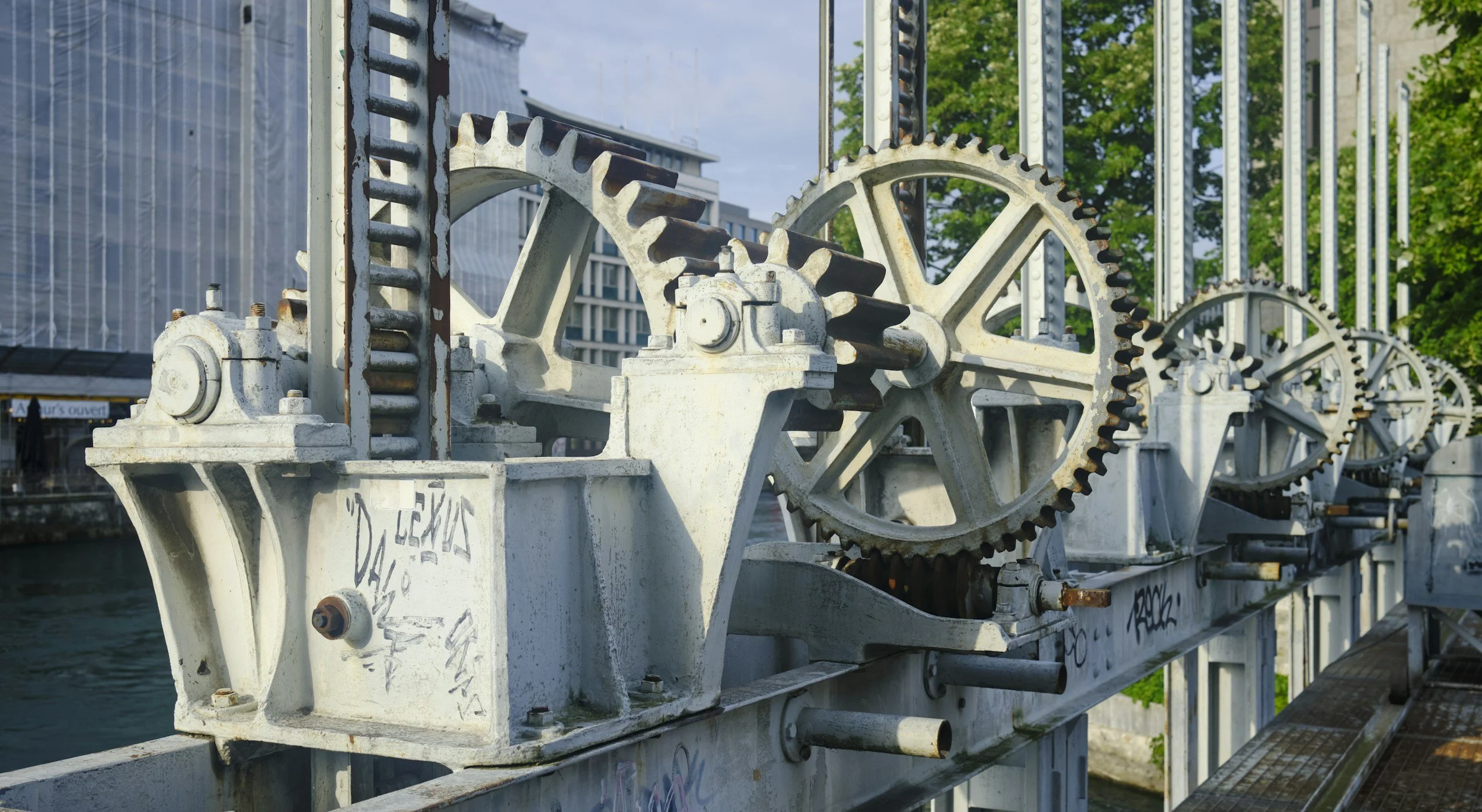 Close-up of large metal gears and mechanical components on a bridge near water in an urban area, with trees and buildings in the background.