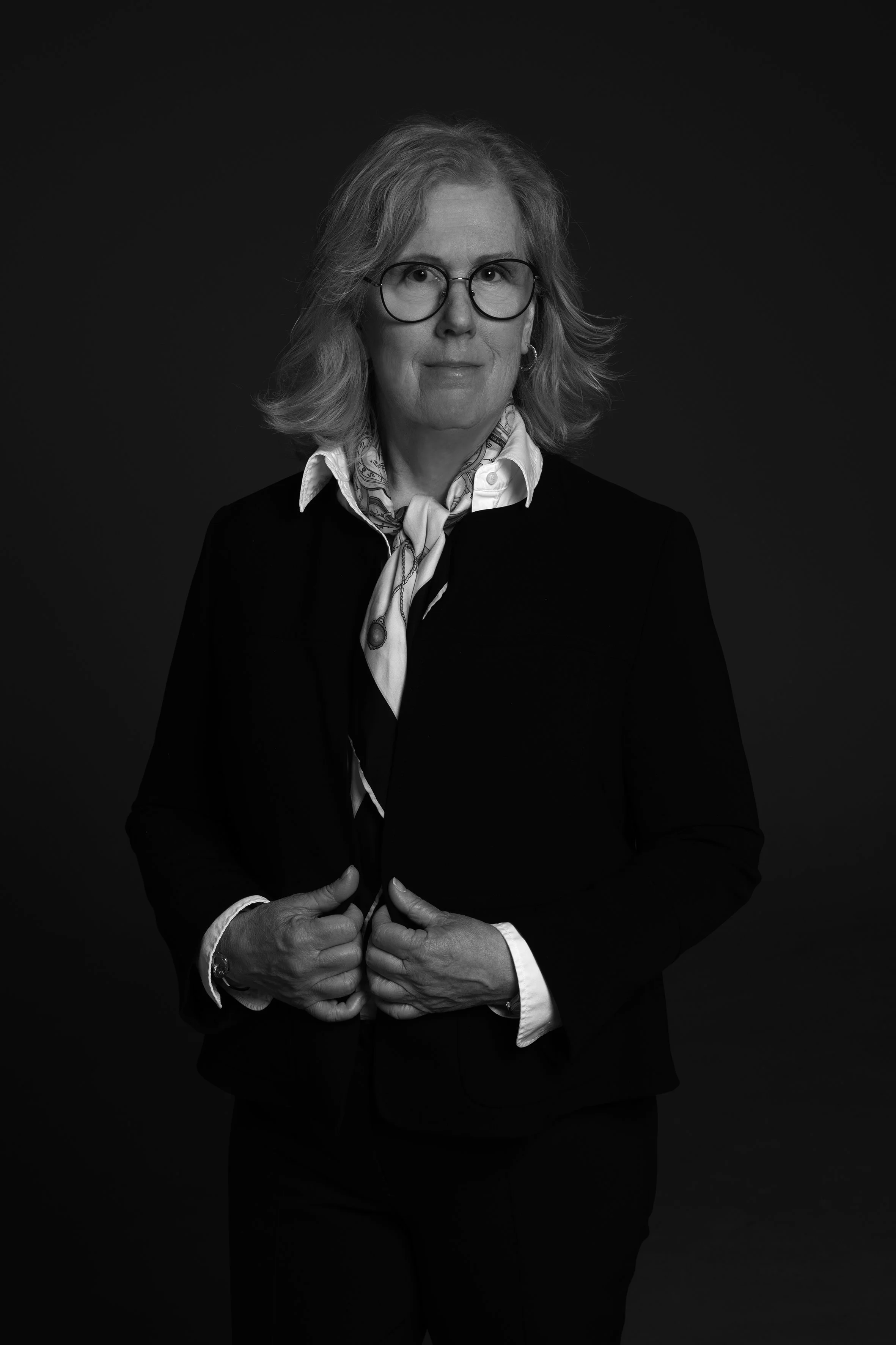En svartvit portrait av en kvinna med glasögon, vit skjorta och scarf, klädd i svart kostym. Hon står mot en mörk bakgrund.