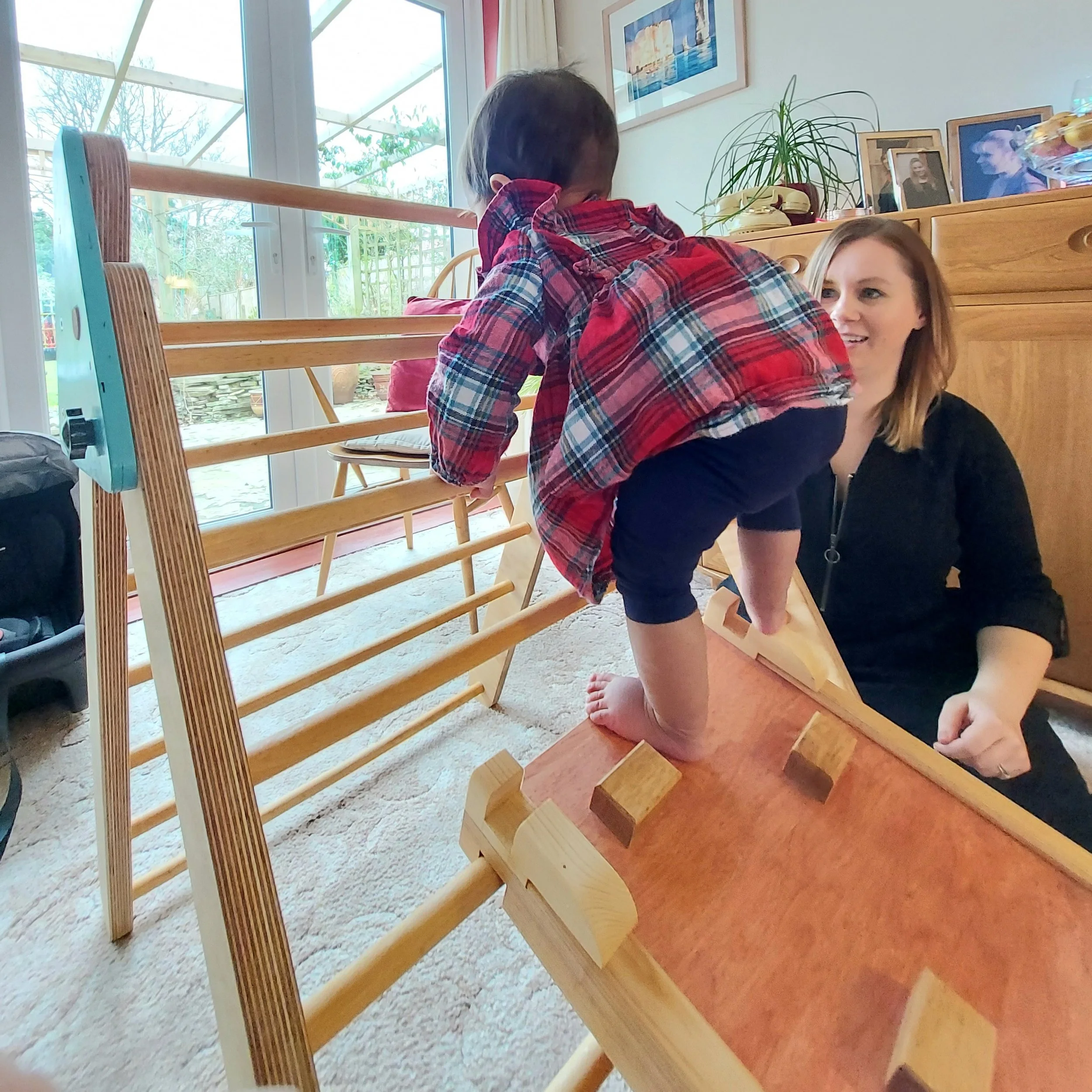 A young boy in a plaid shirt climbing on a wooden ramp with a woman indoors near a glass door viewing a garden.