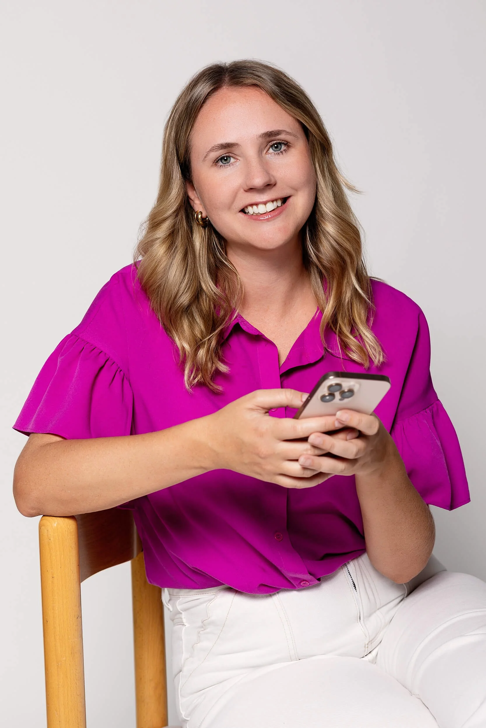 A smiling woman with wavy blonde hair, wearing a bright pink blouse and white pants, sitting on a wooden chair and holding a smartphone.