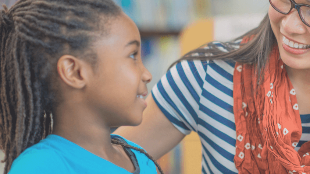 A young girl with braided hair smiling and looking at a woman with glasses, both engaging in a conversation.