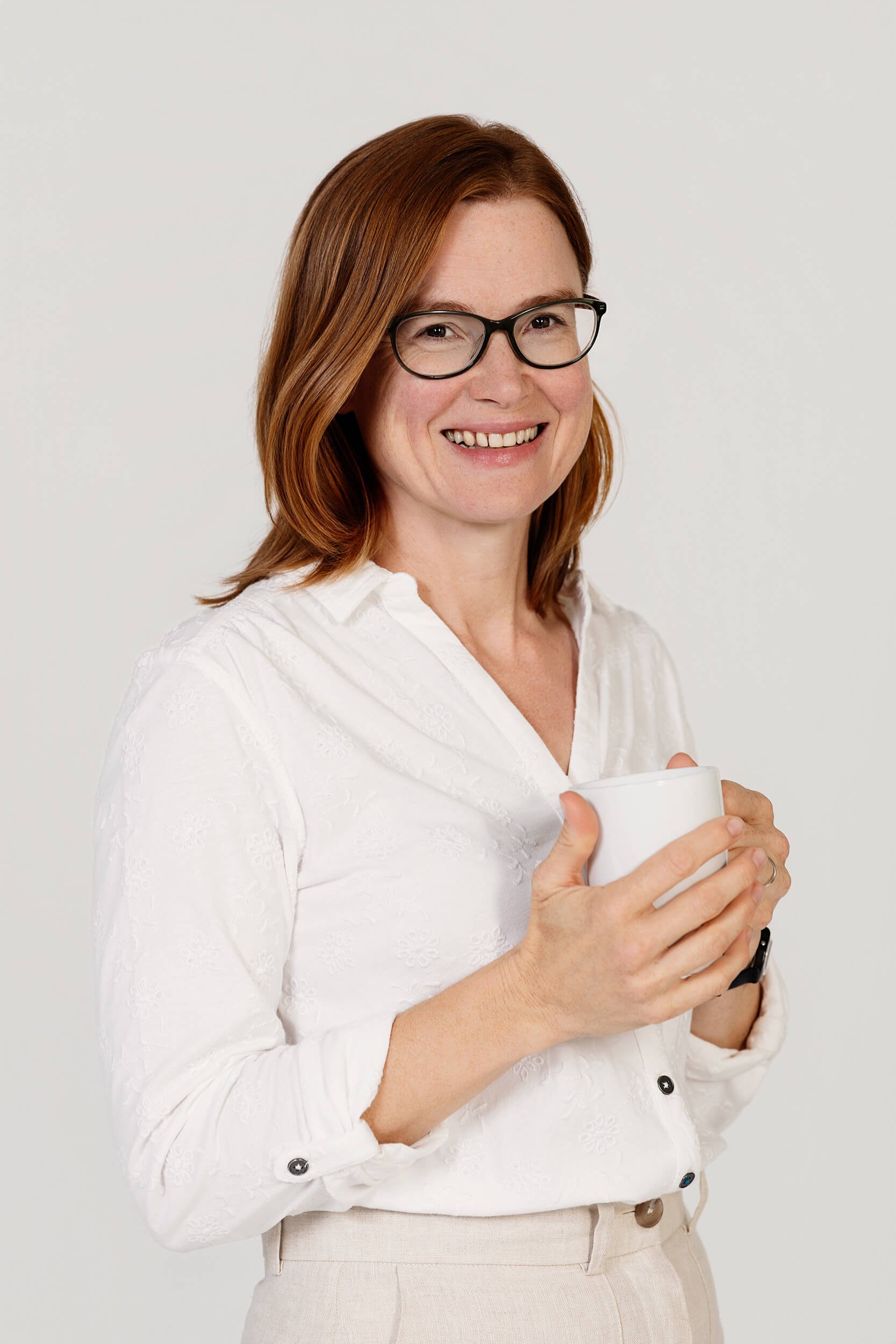 Image of a woman with red hair, glasses, wearing a white blouse, holding a white mug and smiling.