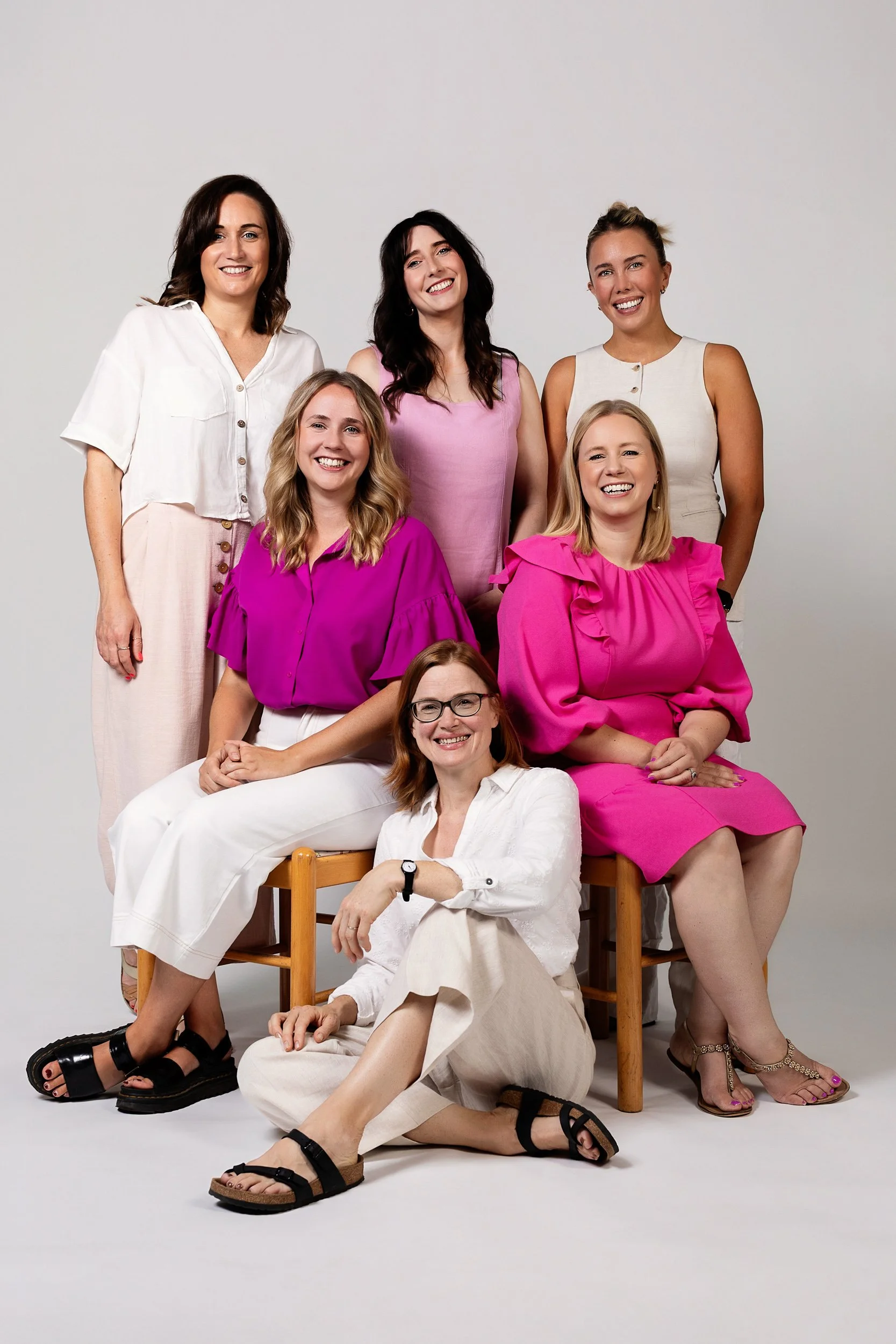 Group of six women smiling and posing together in a studio against a neutral background, some seated and some standing, dressed in bright and neutral colored summer outfits.