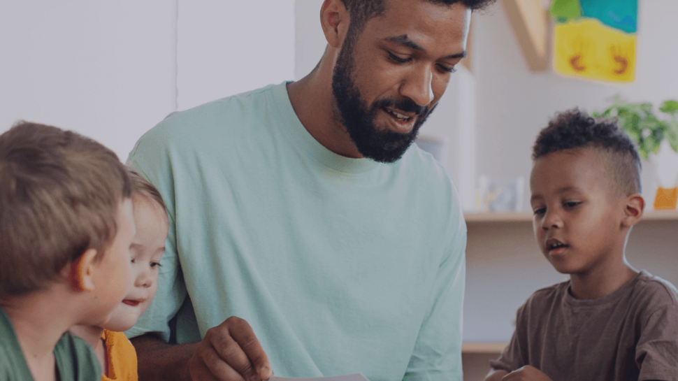 A man with a beard and light green shirt engaging with three young children in a brightly lit room, possibly a classroom or playroom, with children looking at him attentively.