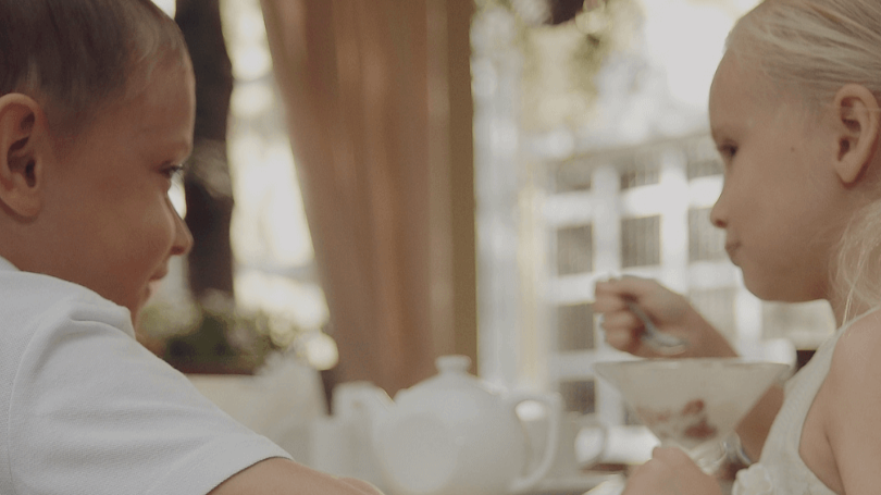 Two children eating breakfast at a table with a bowl and teapot, sitting indoors near a window.