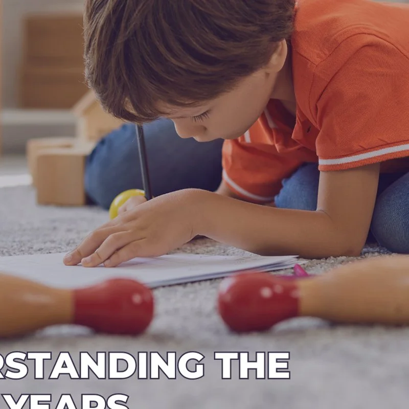 Child drawing or writing on a piece of paper while lying on the carpeted floor, with wooden toys around him.