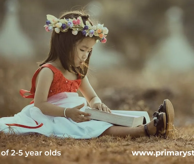 A young girl wearing a red dress and a flower crown, sitting on the ground outdoors, reading a large book.