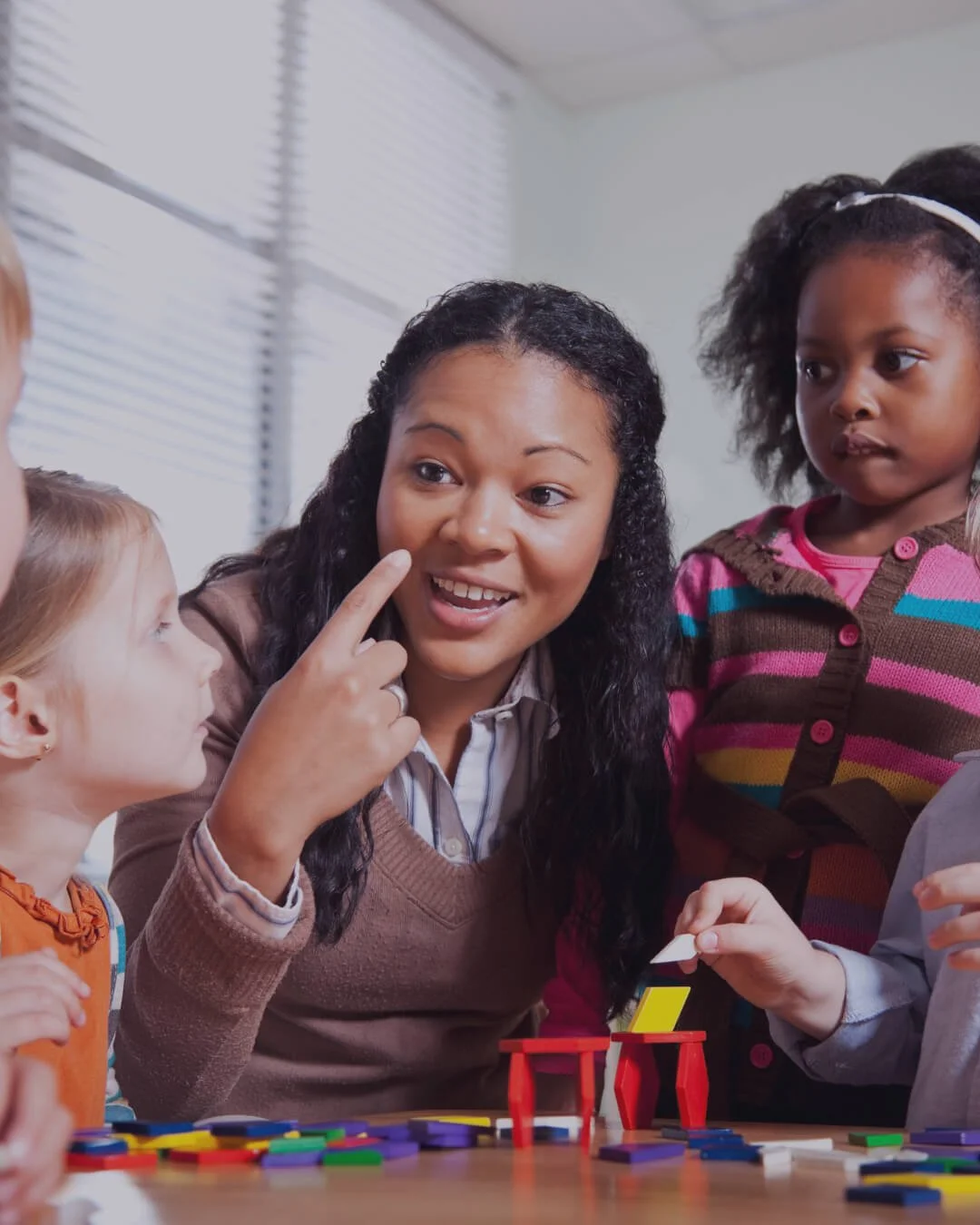 A woman pointing at her face while children listen during a classroom activity with colorful game pieces on the table.