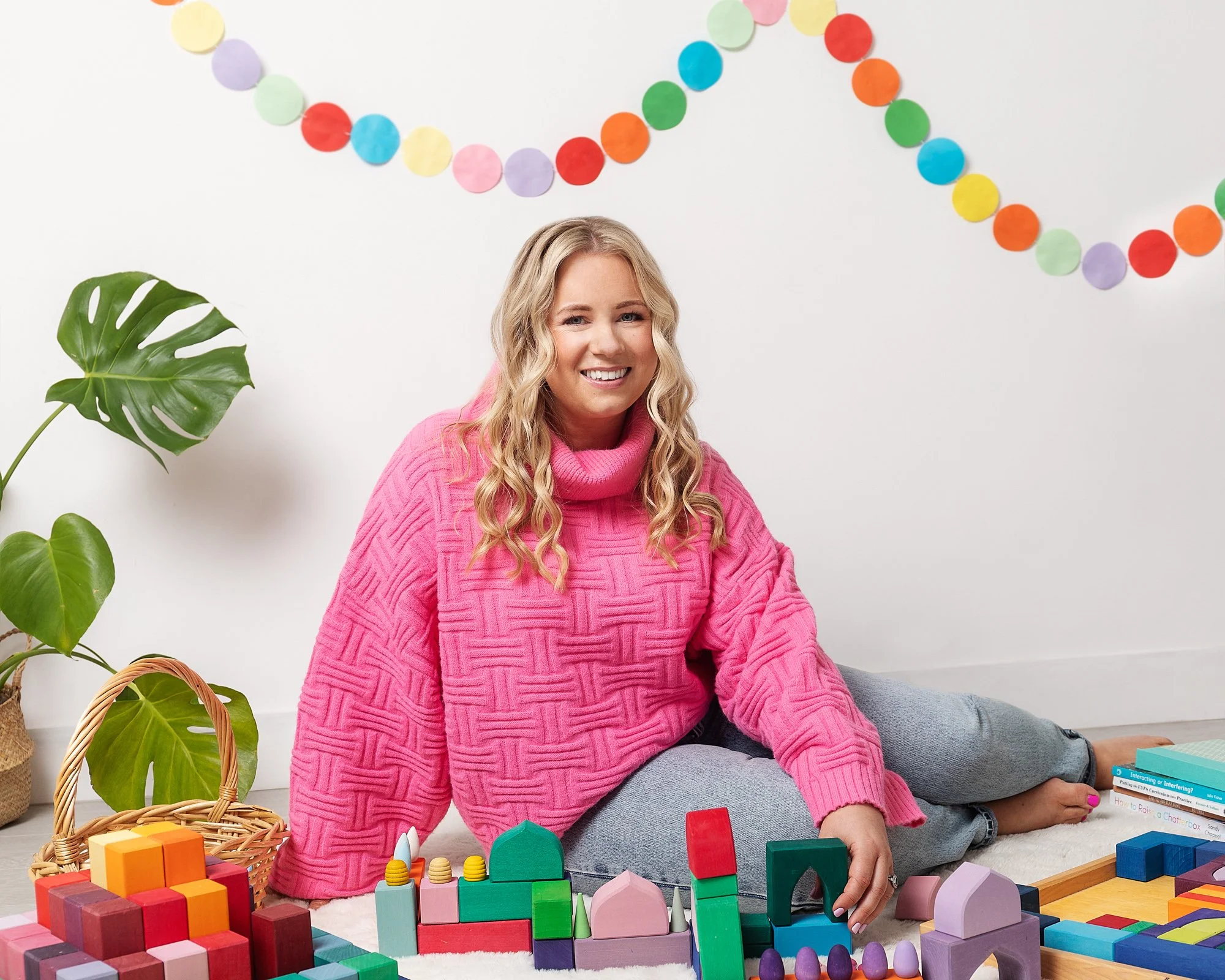 A woman with blonde curly hair sitting on the floor, smiling, wearing a pink sweater and gray pants, surrounded by colorful wooden blocks and toys, with a large green leaf plant and a paper chain decoration on the wall in the background.