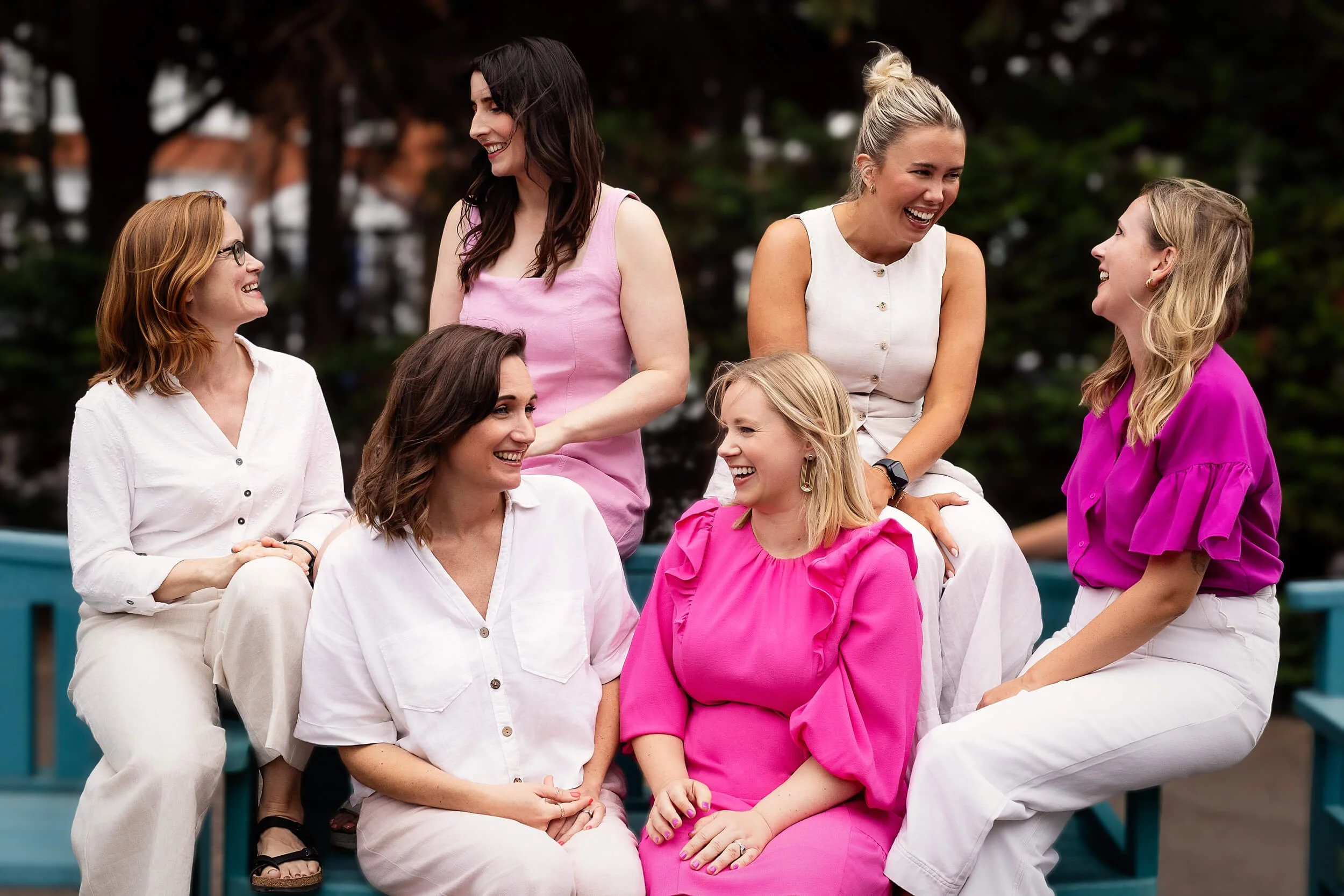 Six women sitting on a blue bench outdoors, engaged in conversation and laughter, with trees in the background.