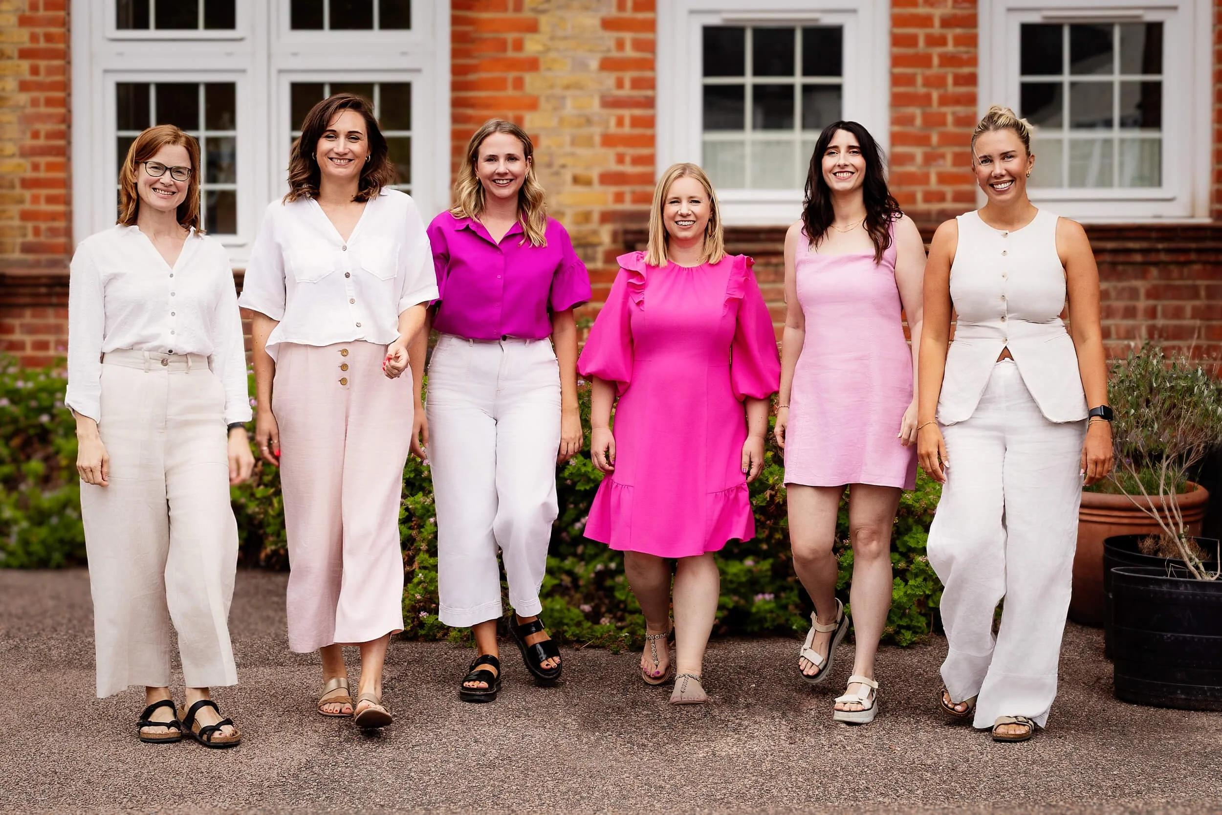 Six women walking together outside in front of a brick house, smiling, wearing casual summer clothes in white and pink tones.