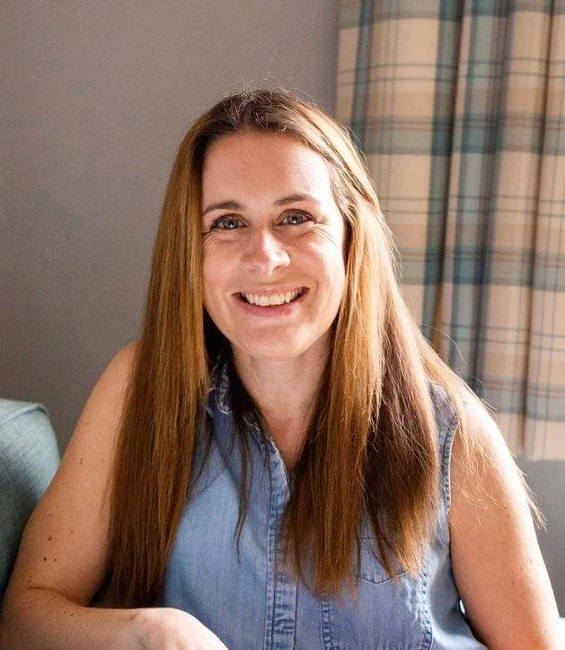 Smiling woman with long reddish-brown hair wearing a sleeveless denim shirt sitting in a room with a window and plaid curtains.
