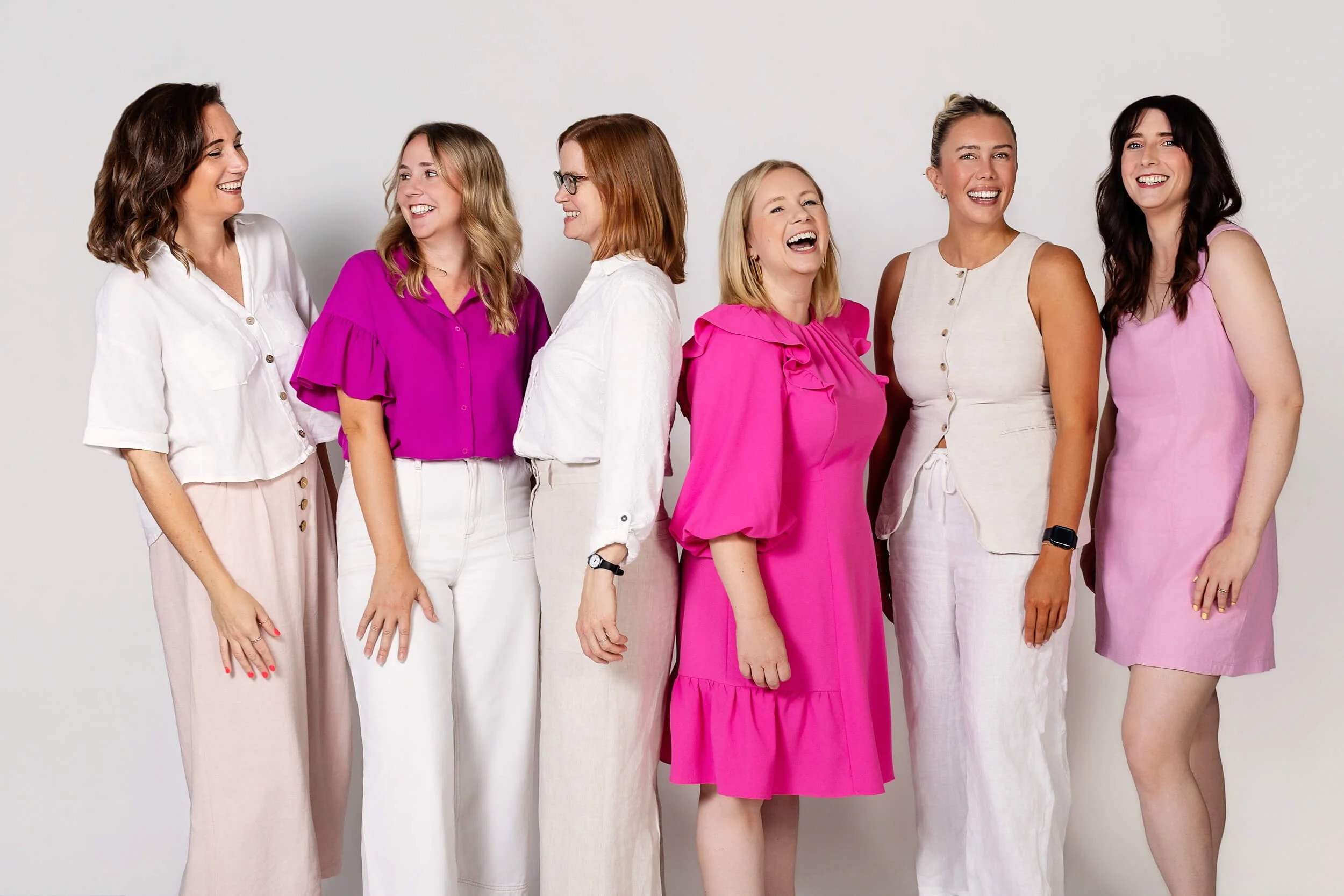 Six women standing in a row against a white background, laughing and smiling.
