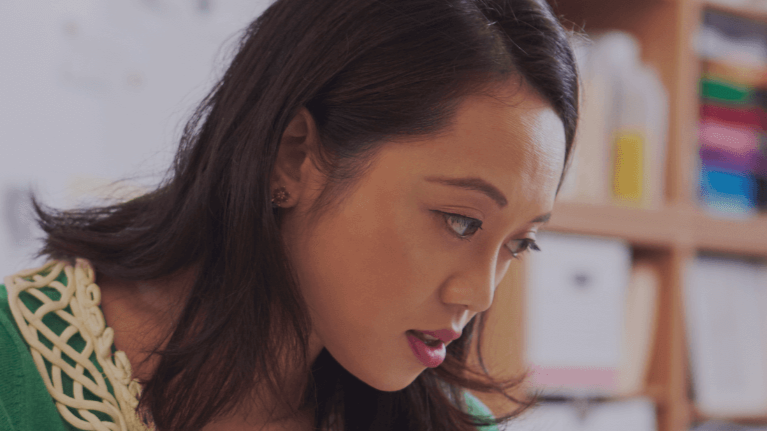 Close-up of a young woman with dark hair looking down, possibly reading or working, with books and shelves in the background.