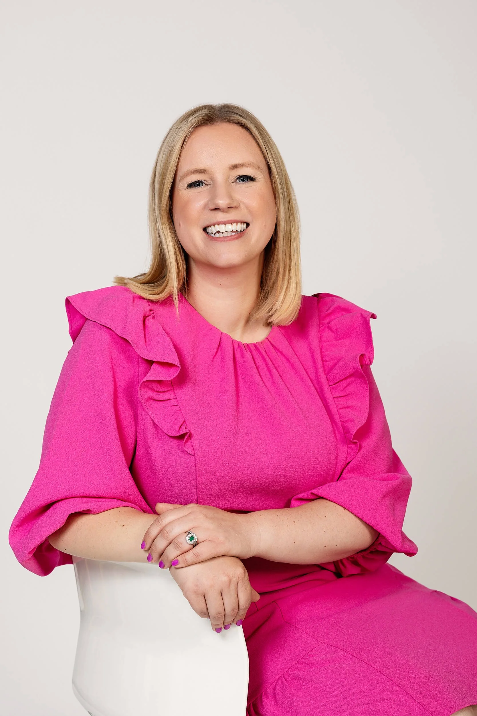 A smiling woman with blonde hair in a pink dress with ruffled shoulders sitting on a white chair against a plain white background.