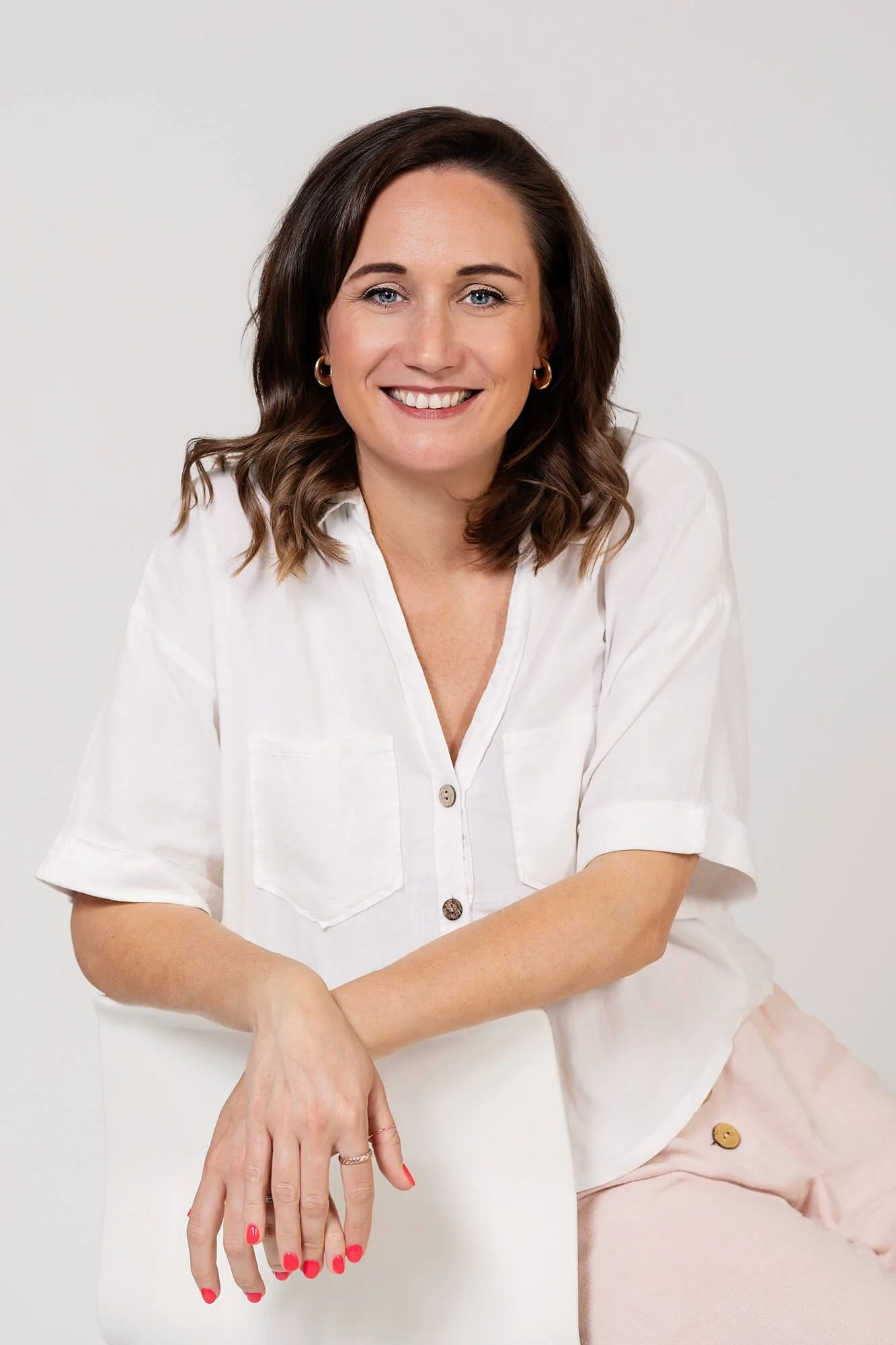 A woman with shoulder-length wavy brown hair, wearing a white button-up shirt and pink pants, smiling and sitting on a white chair against a plain light-colored background.