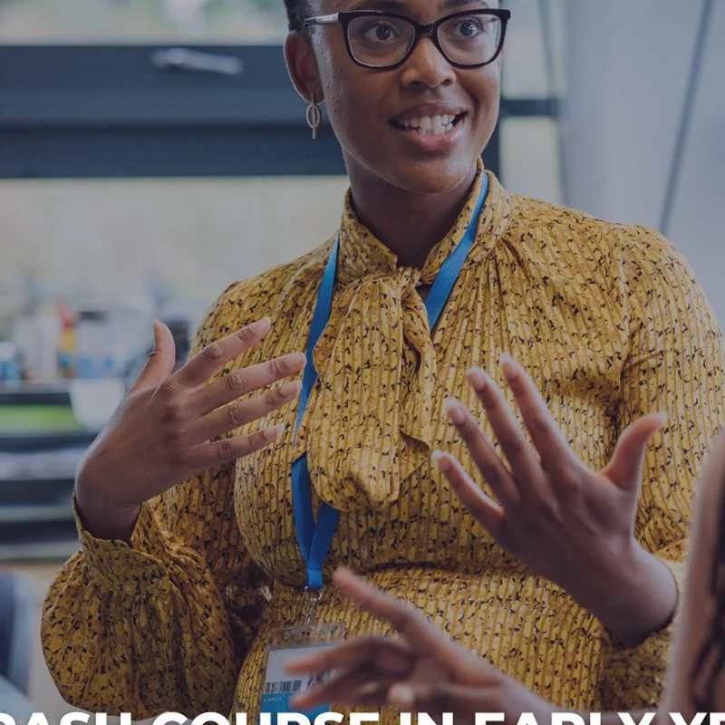 A woman in glasses wearing a yellow patterned blouse and a blue lanyard is talking and gesturing with her hands in an indoor setting, possibly an office.