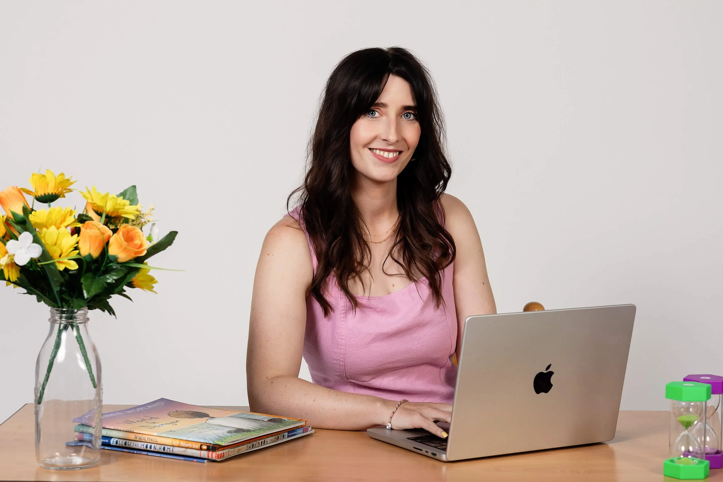 A woman with long dark hair wearing a pink sleeveless top sitting at a desk with a silver MacBook, a bouquet of yellow and orange flowers in a glass vase, a stack of magazines, and colorful hourglasses, against a plain white background.