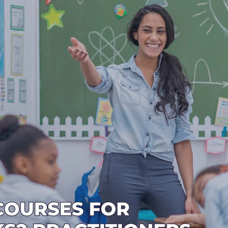 A teacher smiling and pointing in a classroom with students. There are colorful decorations on a green chalkboard with scientific drawings. The students are seated and listening.
