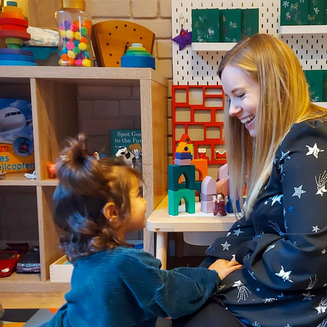 A young girl and a woman are sitting and smiling at each other in a playroom. The girl has curly brown hair with a top knot, and the woman has long blonde hair. There are colorful toys and shelves behind them, including building blocks, plush toys, and game boxes.