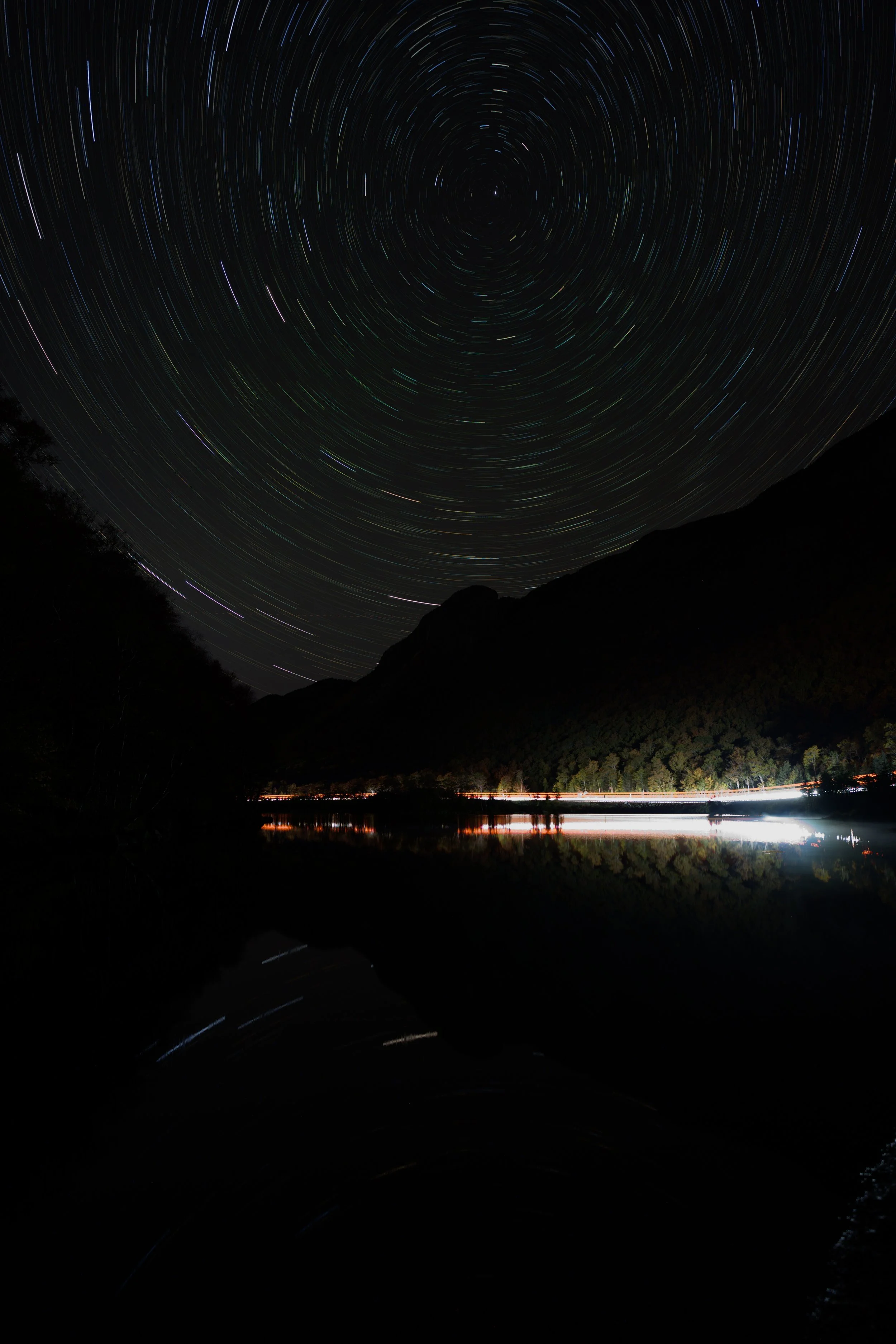 Long exposure photograph of star trails in the night sky over a mountain lake, with the reflection of the stars and mountains visible on the water's surface.