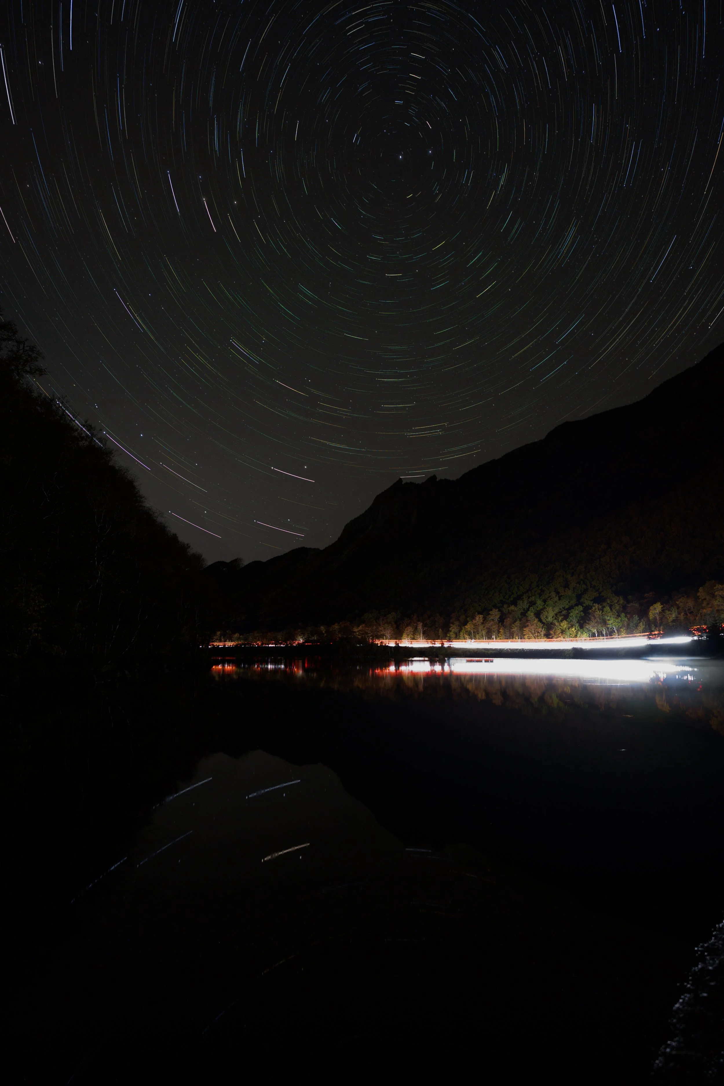 Nighttime landscape with star trails in the sky, mountains, and a reflected calm body of water with light streaks on the horizon.