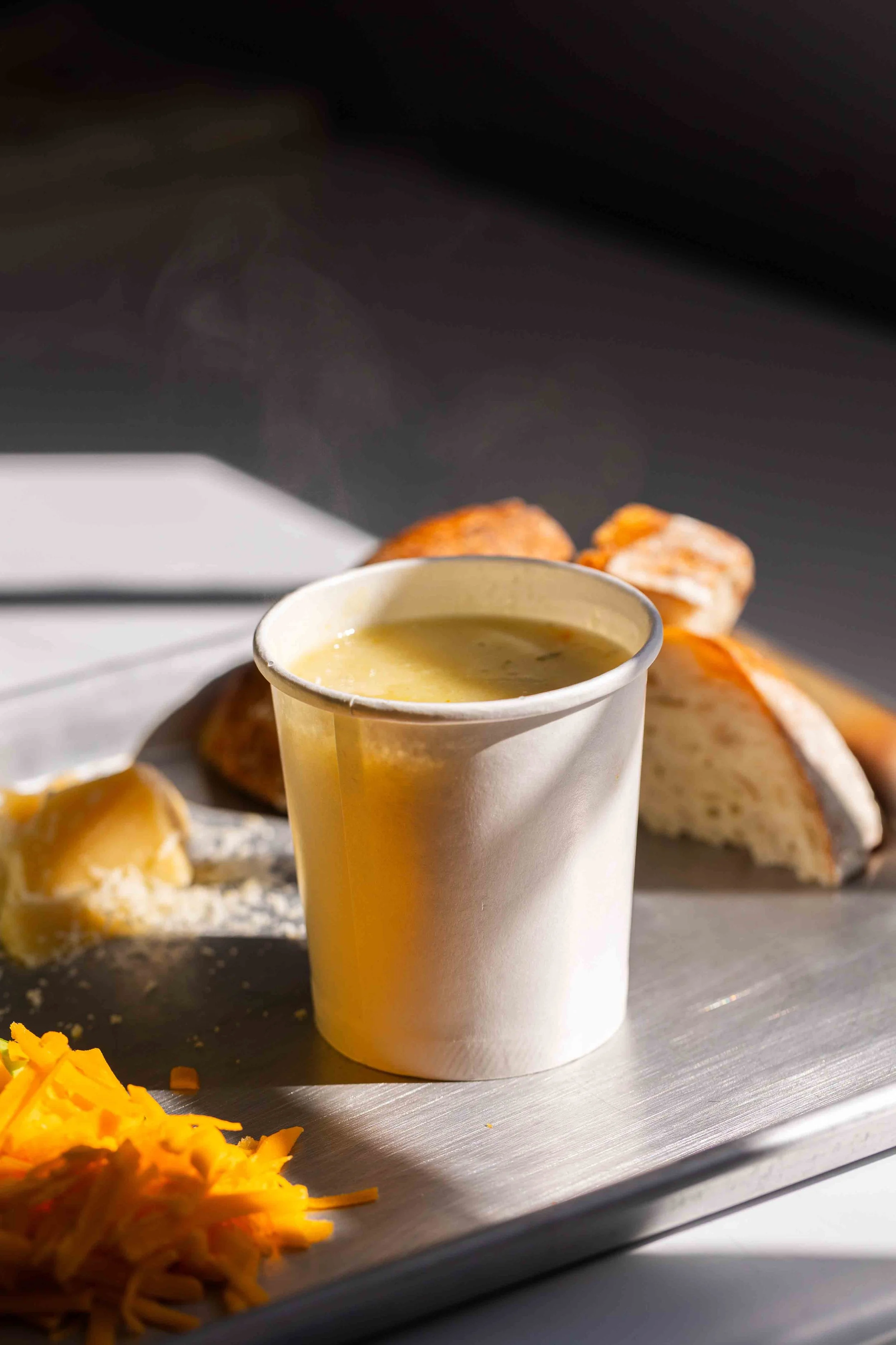 A cup of green soup on a metal tray alongside slices of bread, shredded cheese, a scoop of mashed potatoes, and some biscuits.