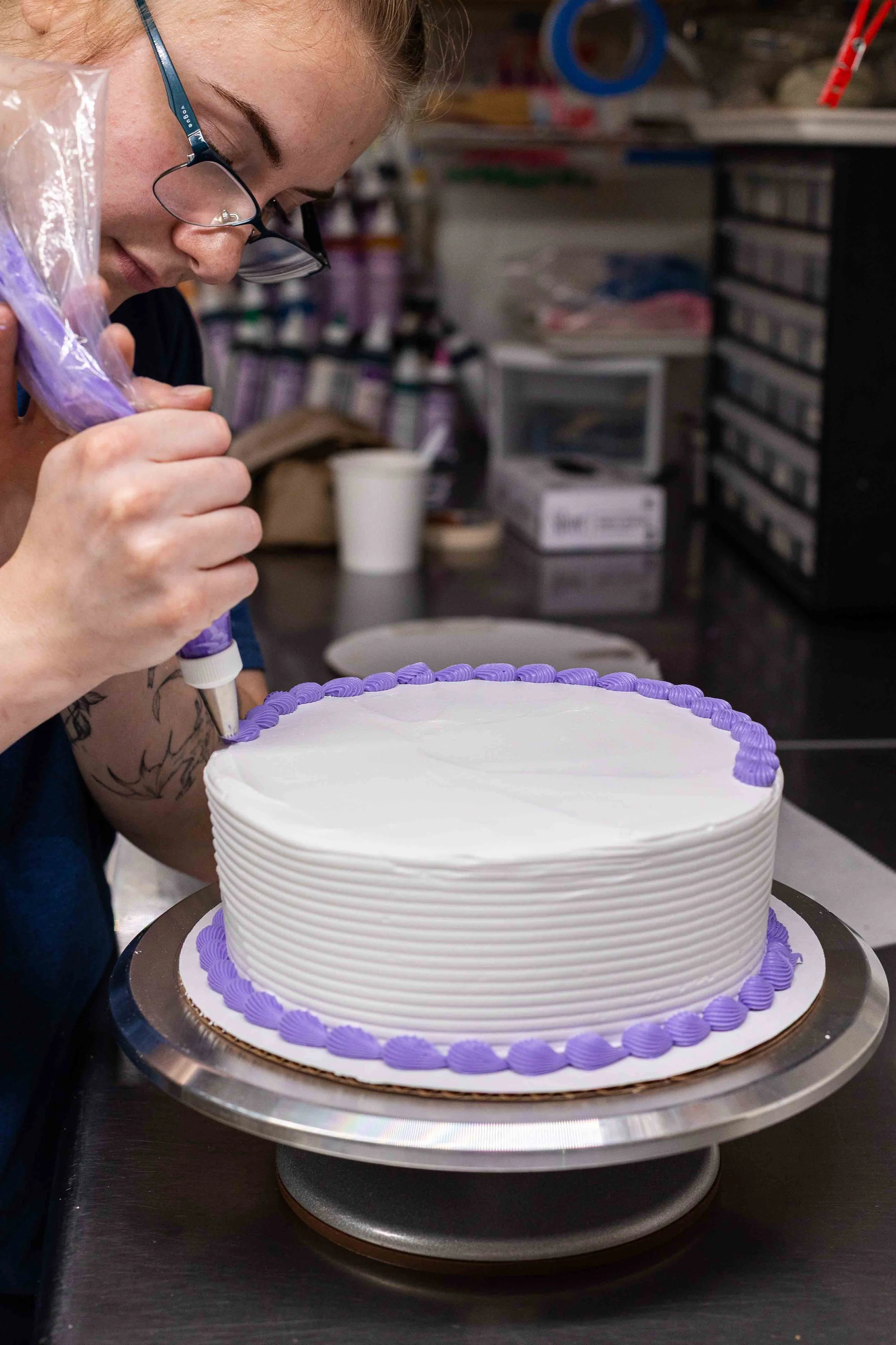 A woman with glasses decorating a white and purple frosted birthday cake with a piping bag in a bakery or kitchen setting.