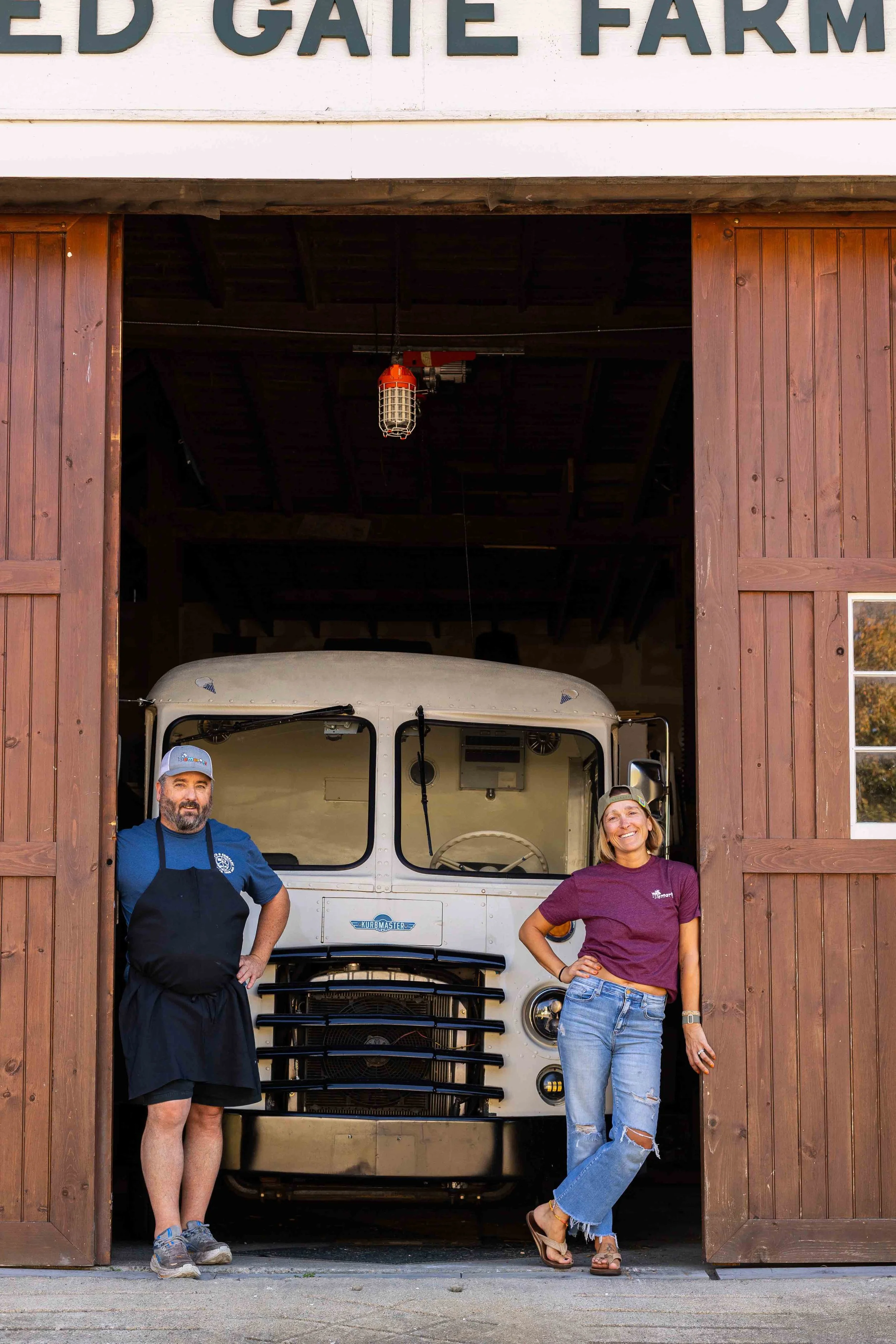 A man and a woman standing in front of a vintage truck inside a wooden barn with an open door. The man is wearing a blue shirt, black apron, and a gray cap, while the woman is dressed in a purple shirt, ripped jeans, and a backward cap, smiling at the camera.